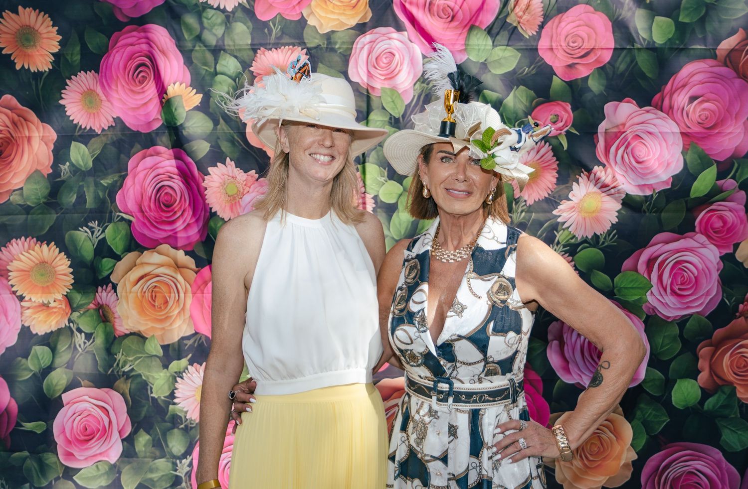 Two women wearing decorative white hats posing together in front of a colorful floral rose wall backdrop.