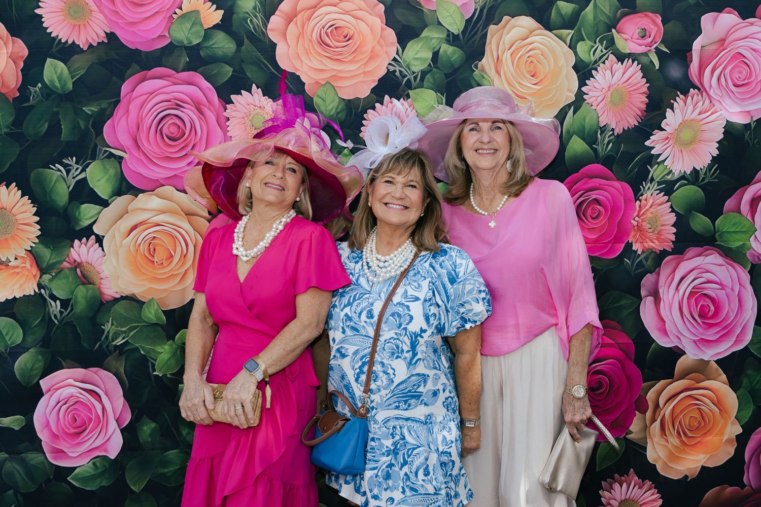 Three women wearing decorative floral hats posing in front of a colorful rose backdrop.