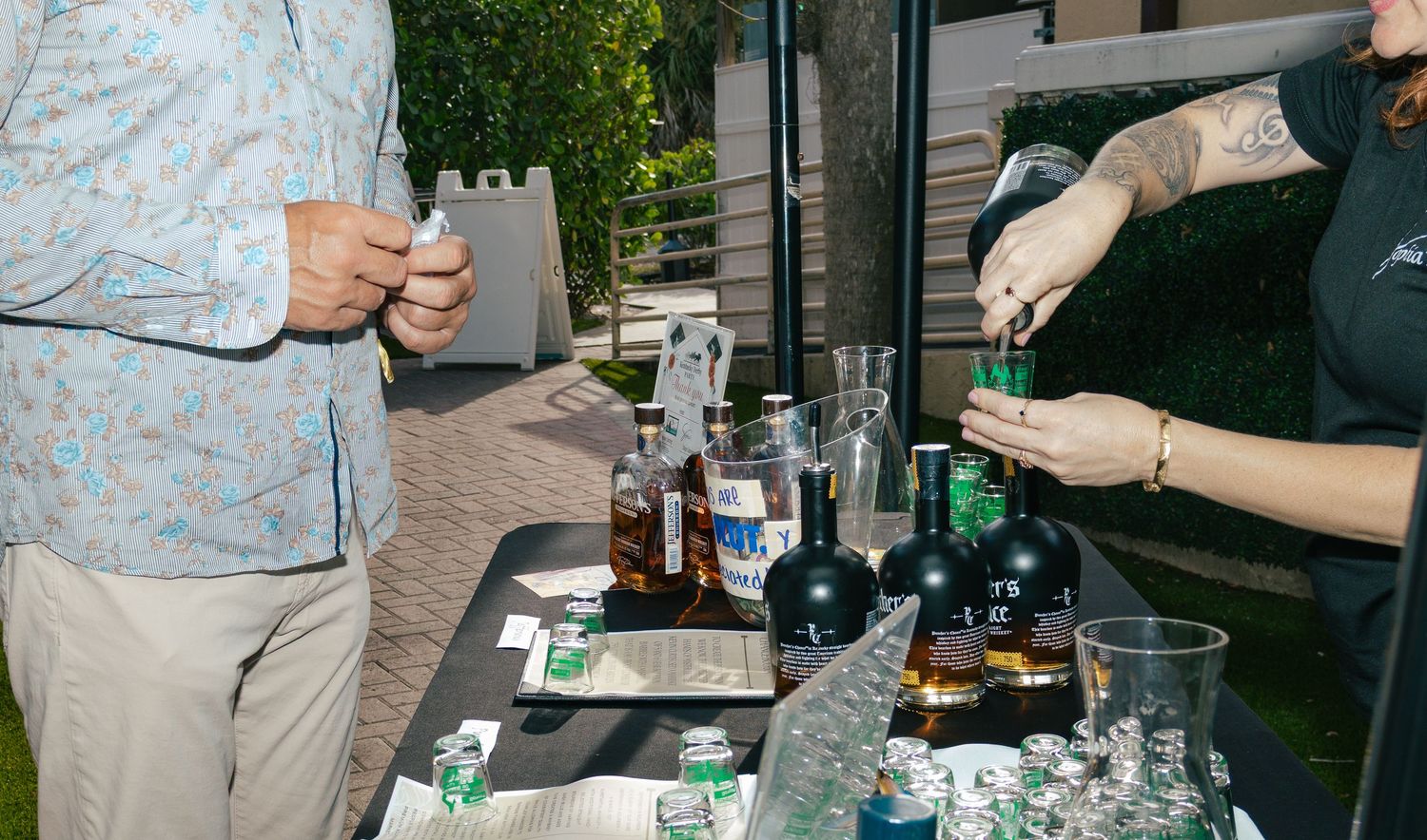Guests enjoy drinks at an outdoor bar setup with whiskey bottles at an evening event.