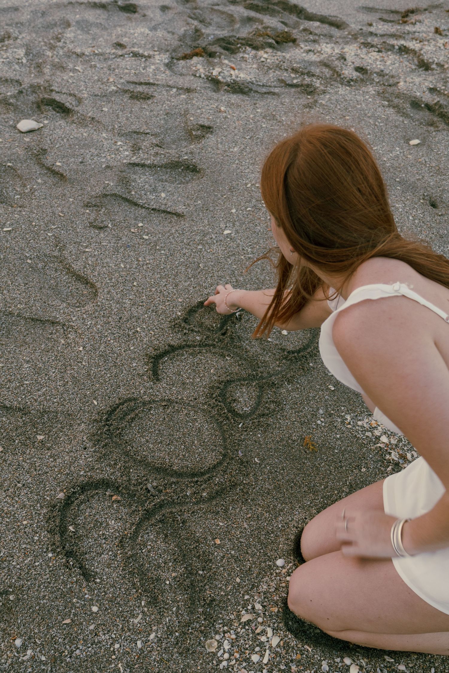 Young woman with auburn hair crouching on dark sand beach writing numbers in the sand.