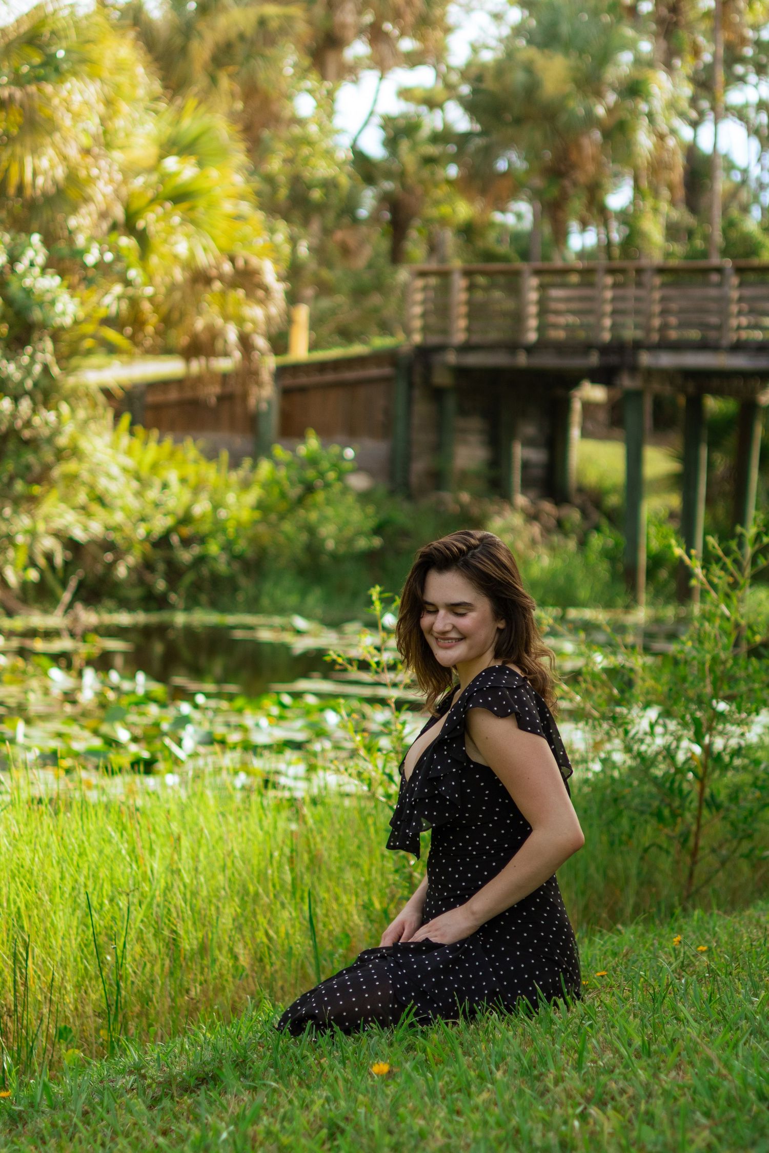 Young woman in black polka dot dress sits on grass beside a scenic pond with wooden bridge.