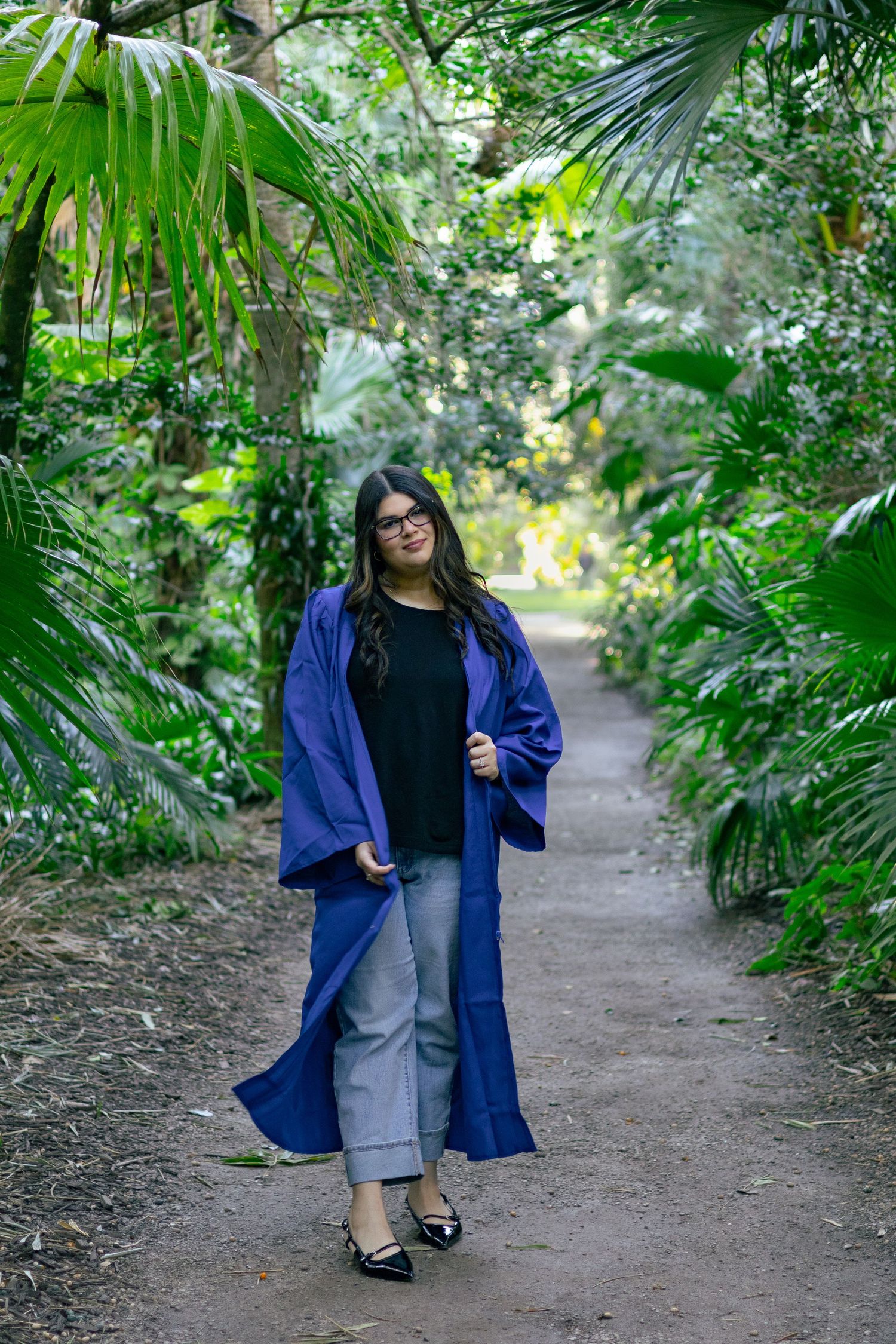 Smiling graduate in open blue graduation gown walking along tropical garden path confidently.