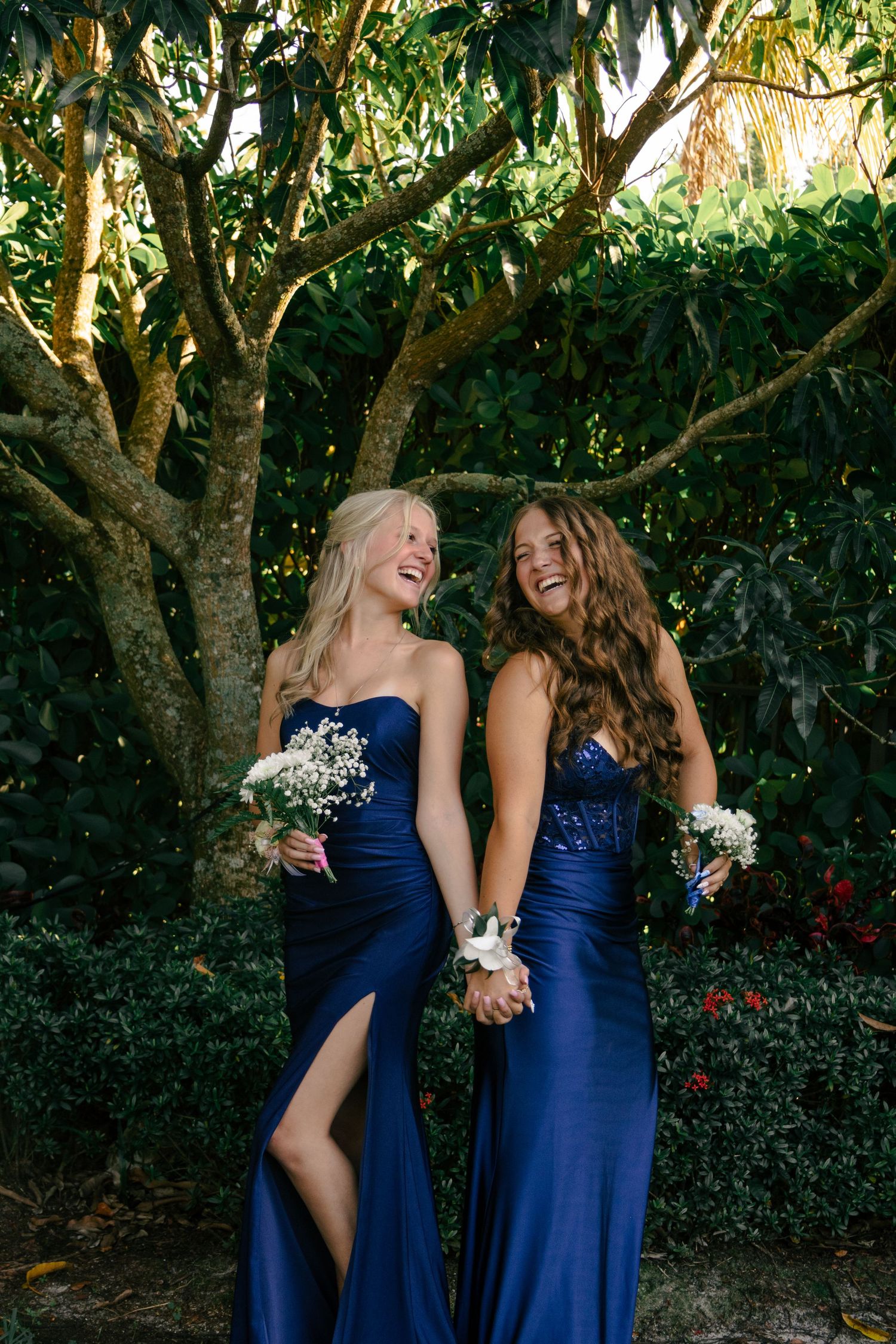 Two laughing young women in matching navy blue prom dresses hold bouquets under a green tree.