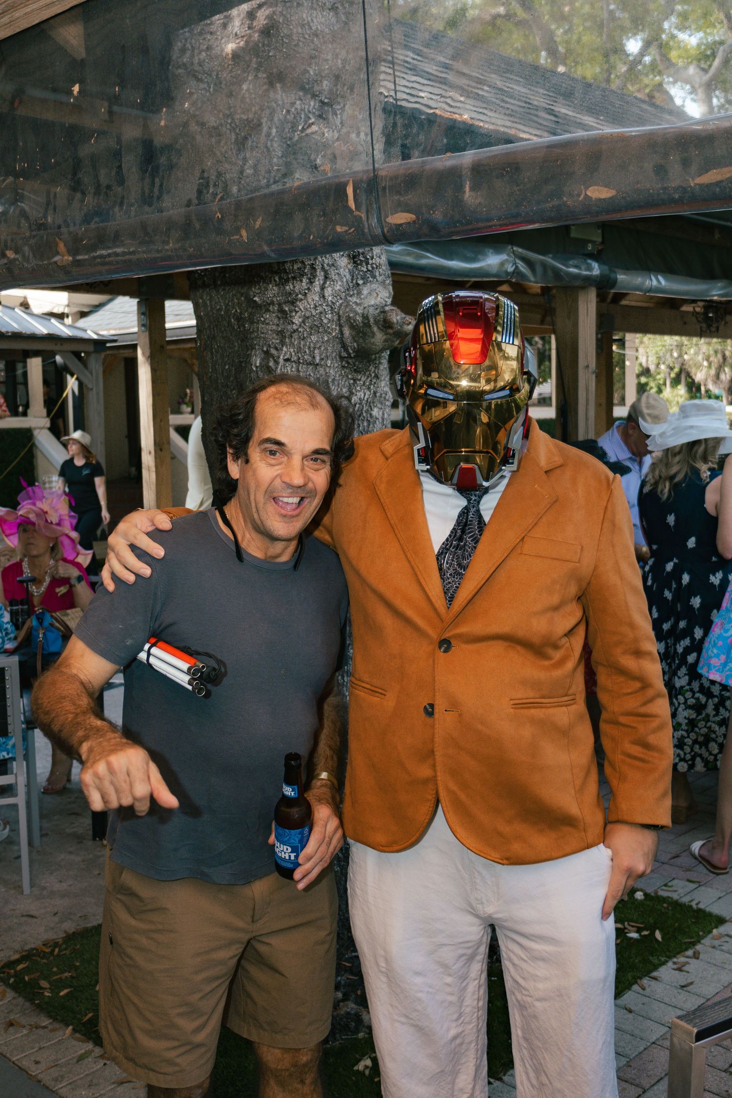 Two men pose outdoors, one wearing an Iron Man helmet and tan blazer at a festive social gathering.