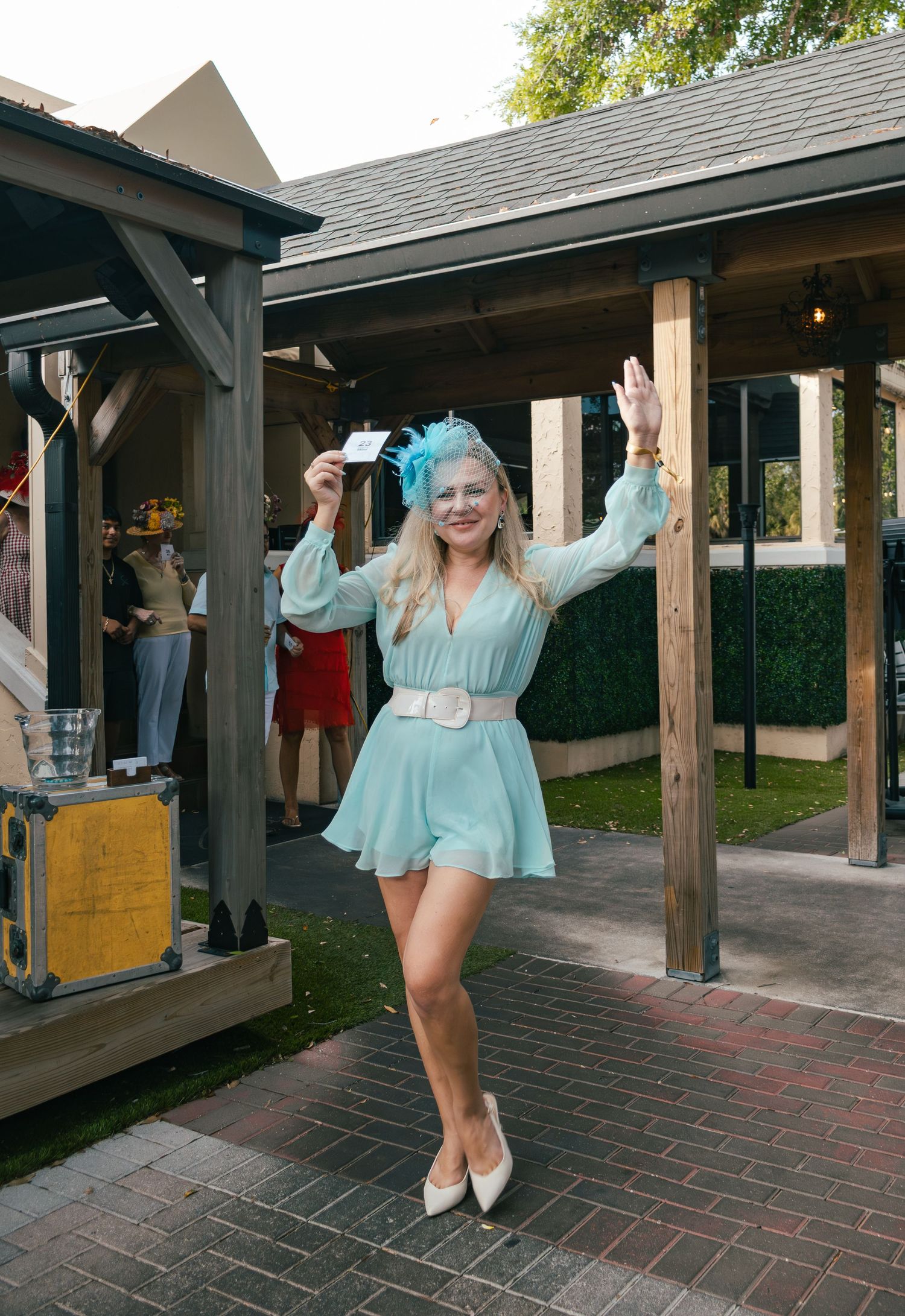 Smiling blonde woman in mint dress and teal fascinator holds card at Kentucky Derby party.
