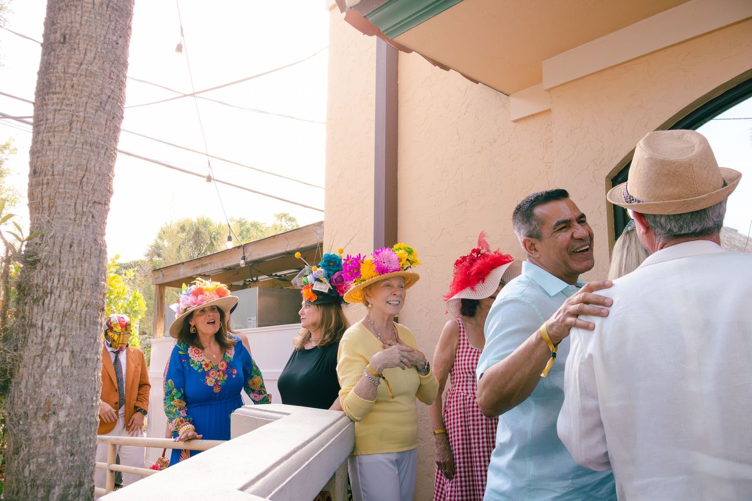 Group of people wearing colorful festive hats socializing on a sunny outdoor patio at a garden party.