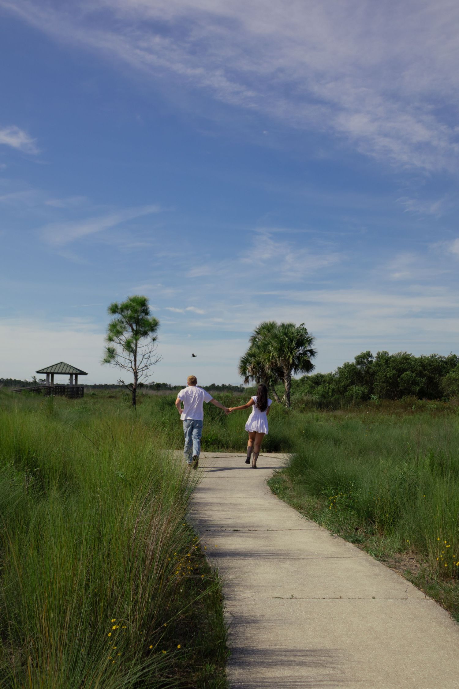 Couple walking away down a concrete path through green marshlands toward a distant gazebo.