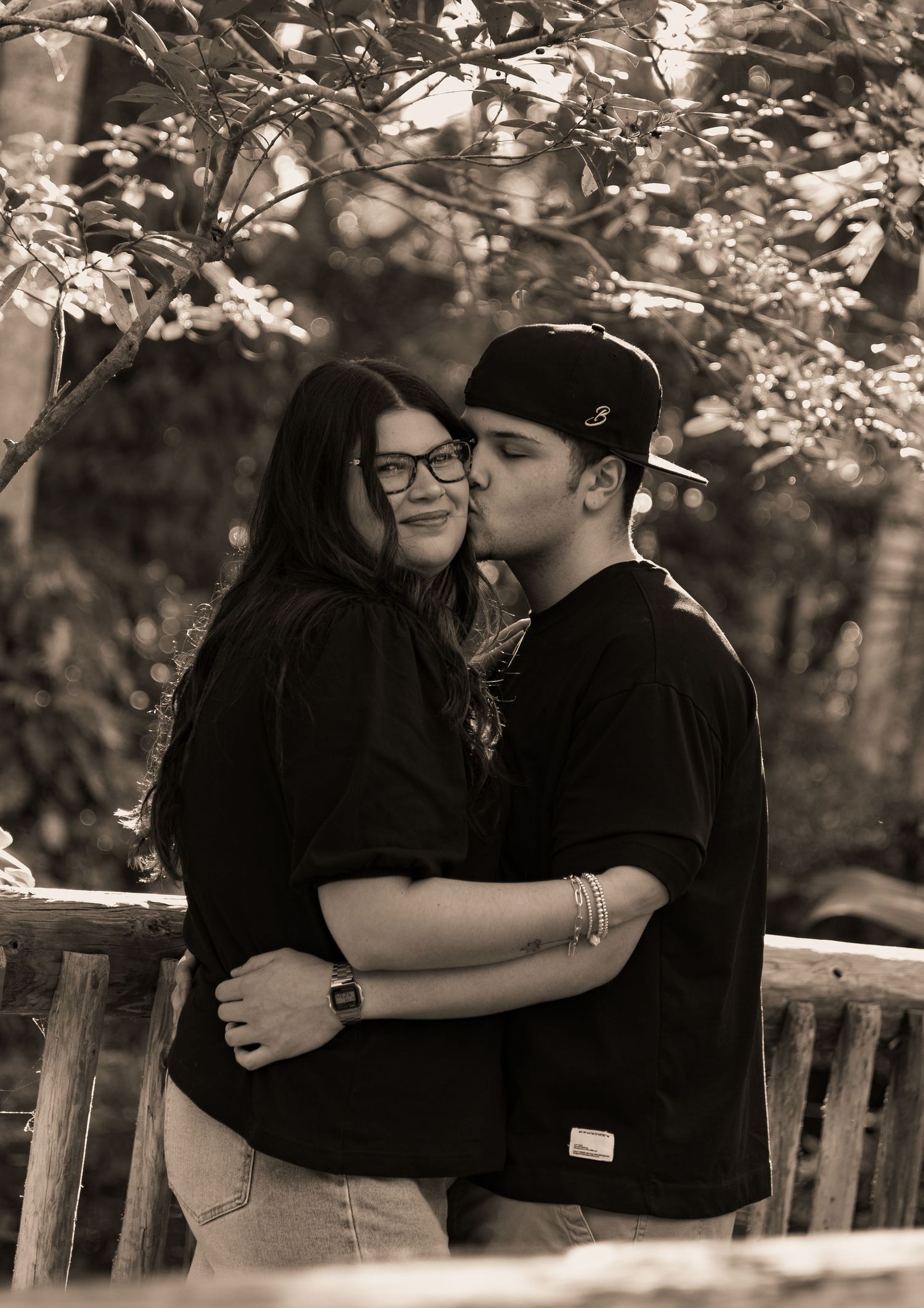 Sepia-toned romantic photo of couple embracing and kissing near wooden fence with bokeh tree background.