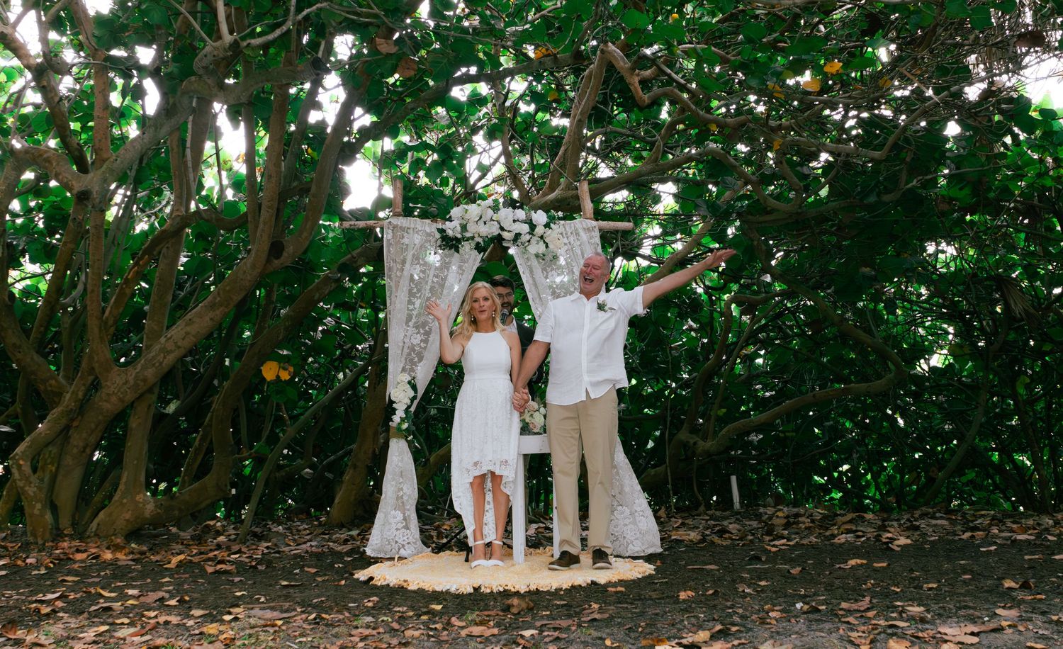 Joyful newlyweds celebrating under floral arch surrounded by lush tropical trees.