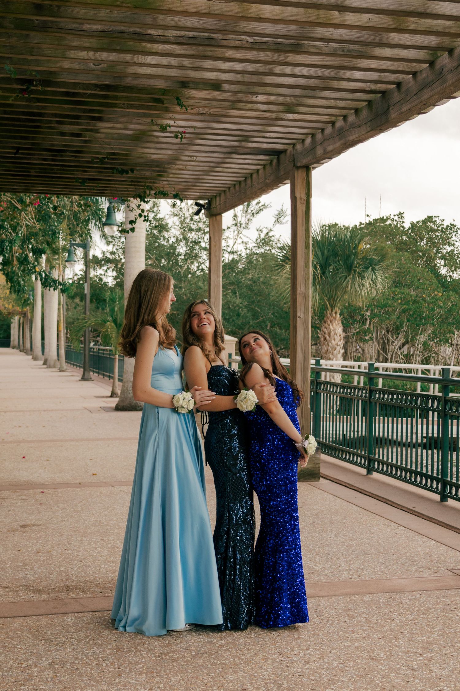 Three girls in elegant blue sequin and satin prom gowns laugh together under an outdoor pergola.
