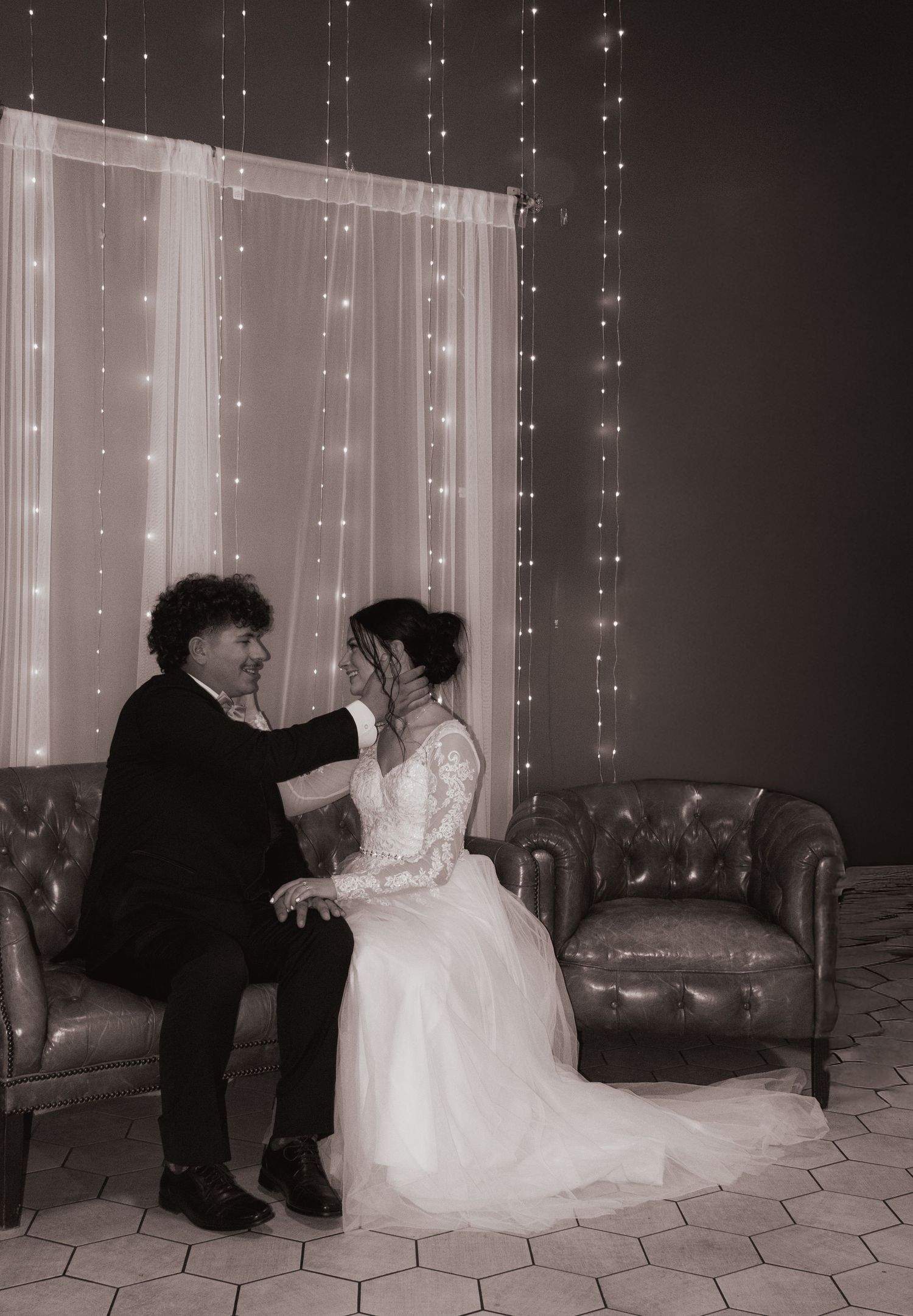 Black and white romantic wedding photo of groom tenderly cupping bride's face while seated on leather chairs with fairy lights.