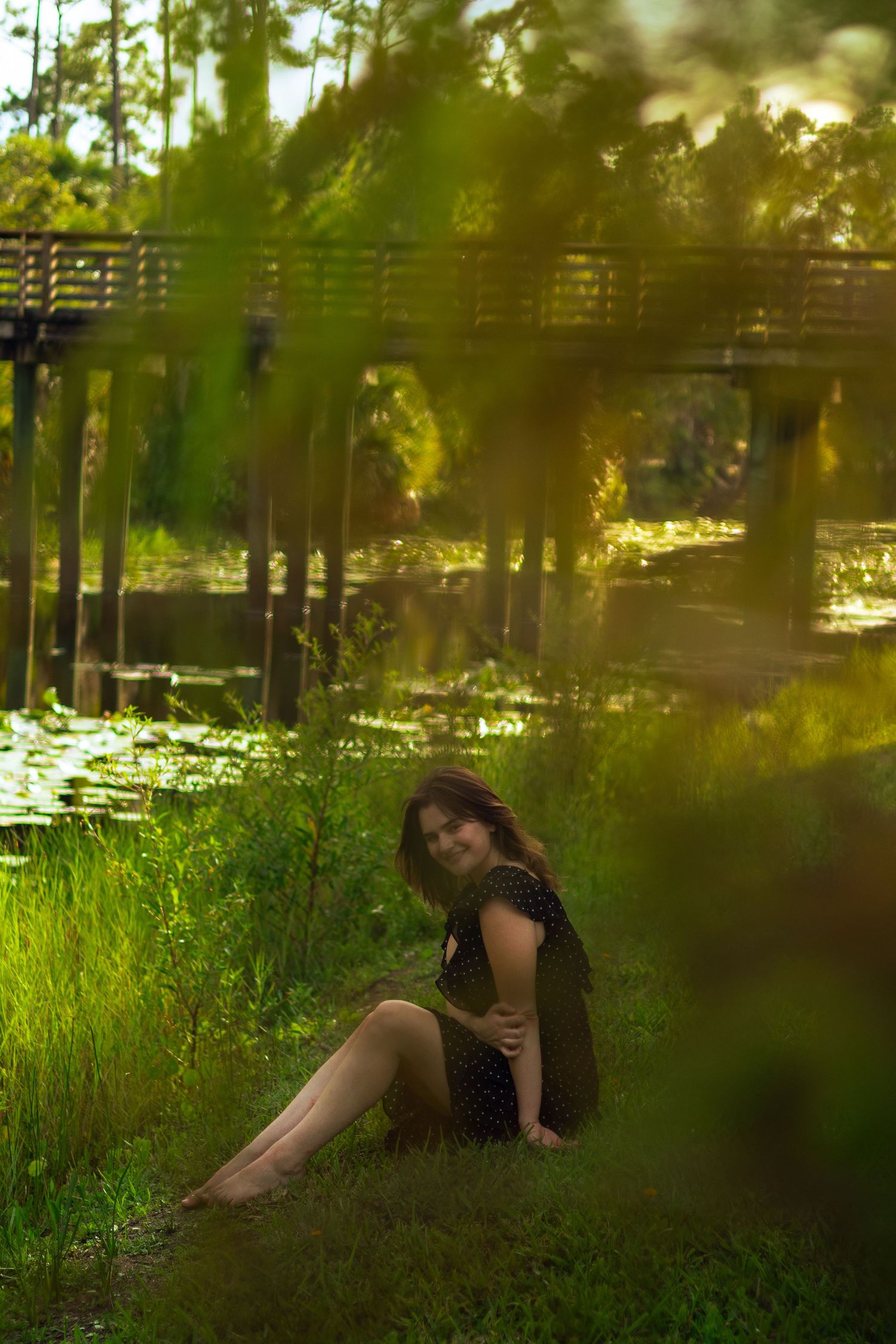 Woman in black polka dot dress sitting barefoot on grass near lily pad pond in golden light.