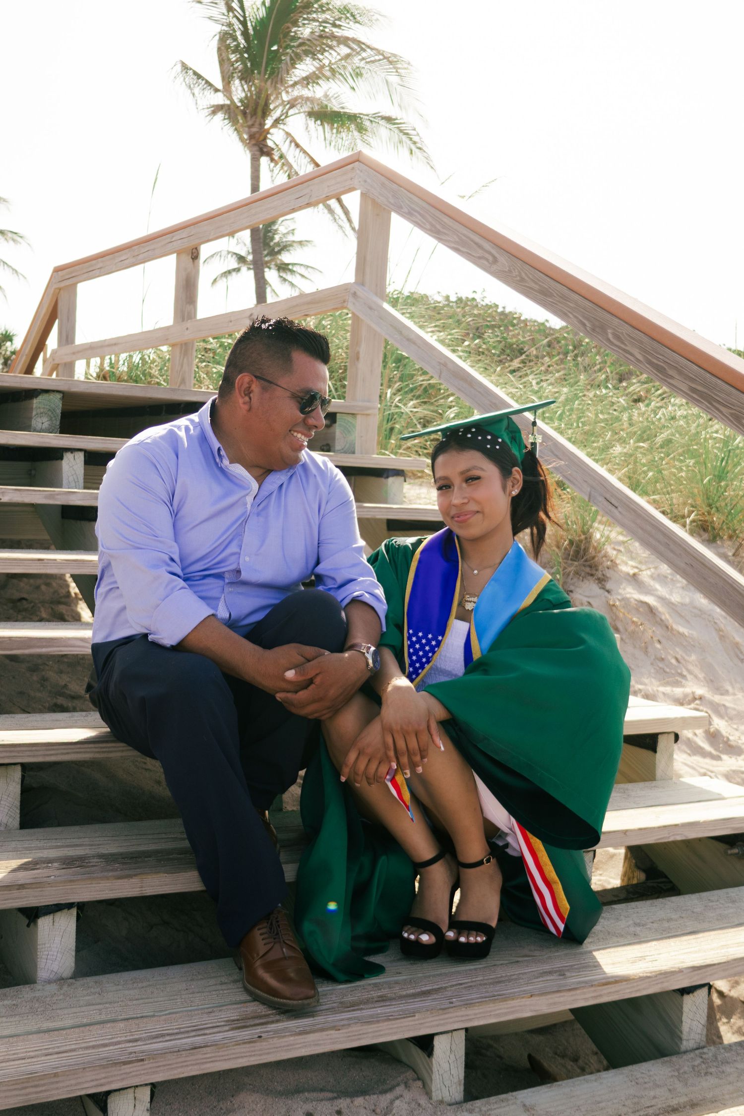 Graduate in green cap and gown sits with father on wooden beach boardwalk stairs.