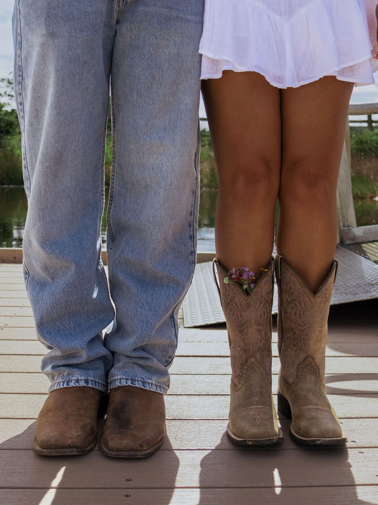 A couple stands on a wooden dock, the woman wearing cowboy boots and a pink dress beside a man in jeans.