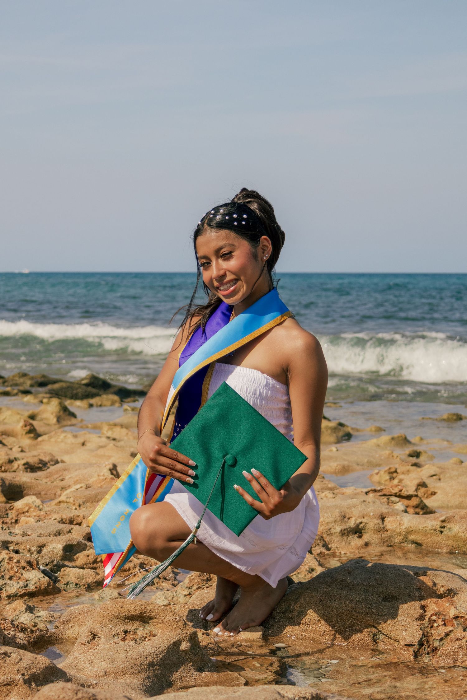 Smiling graduate in white dress holds green cap and sashes while posing on rocky ocean shoreline.