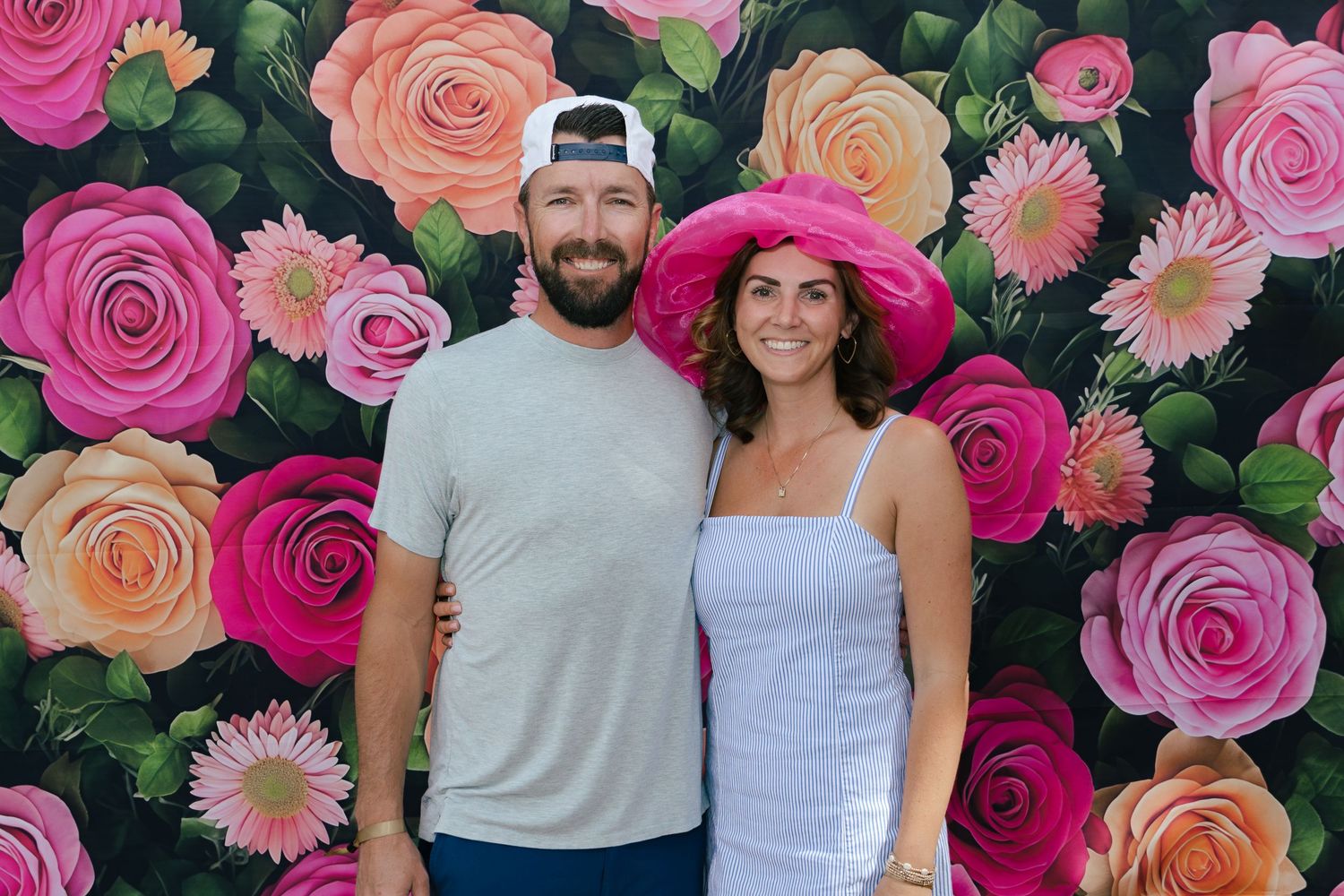 A couple smiles together in front of a bright floral backdrop, woman wearing a pink hat at outdoor event.