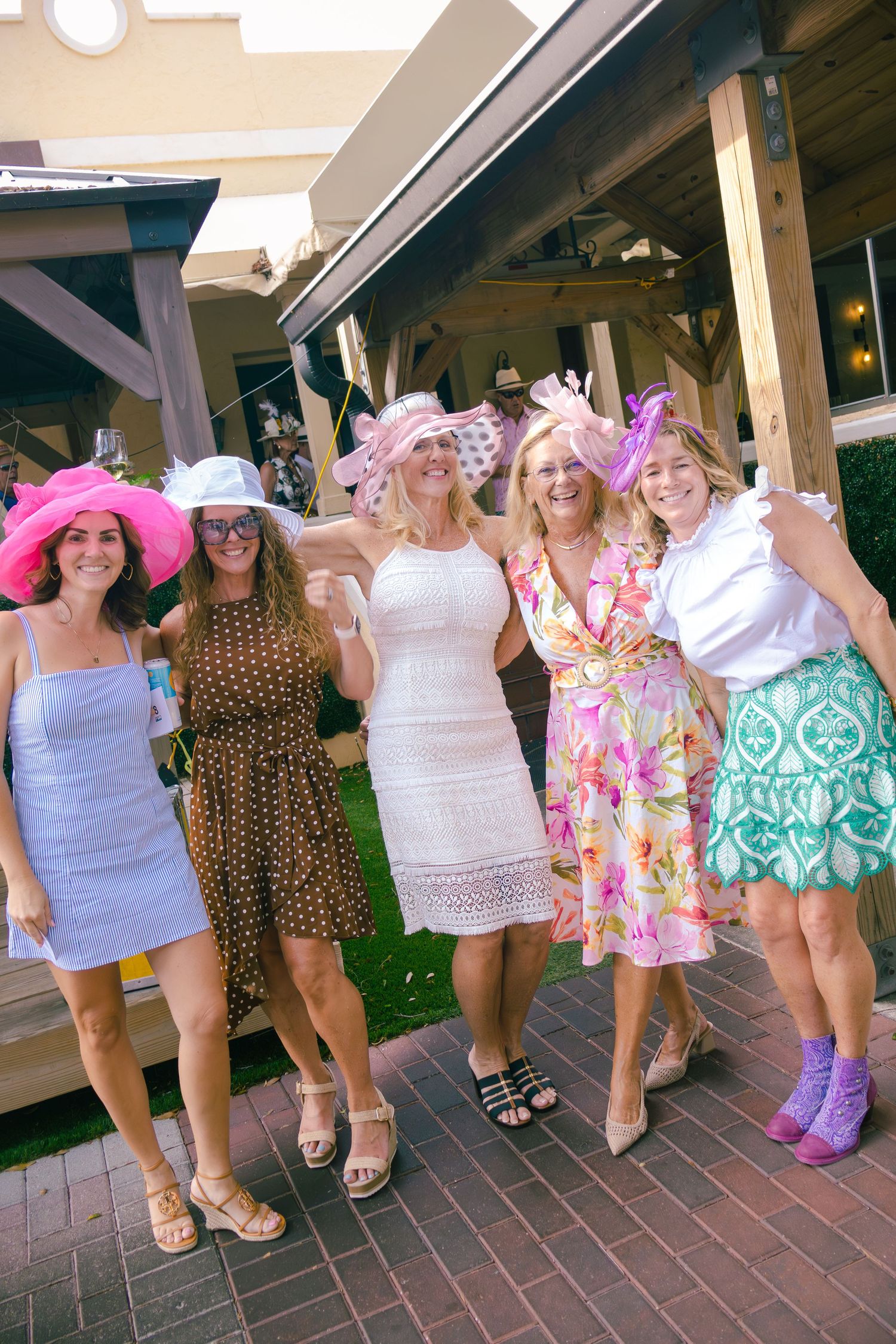 Five women in colorful dresses and decorative hats posing together at outdoor Kentucky Derby party.