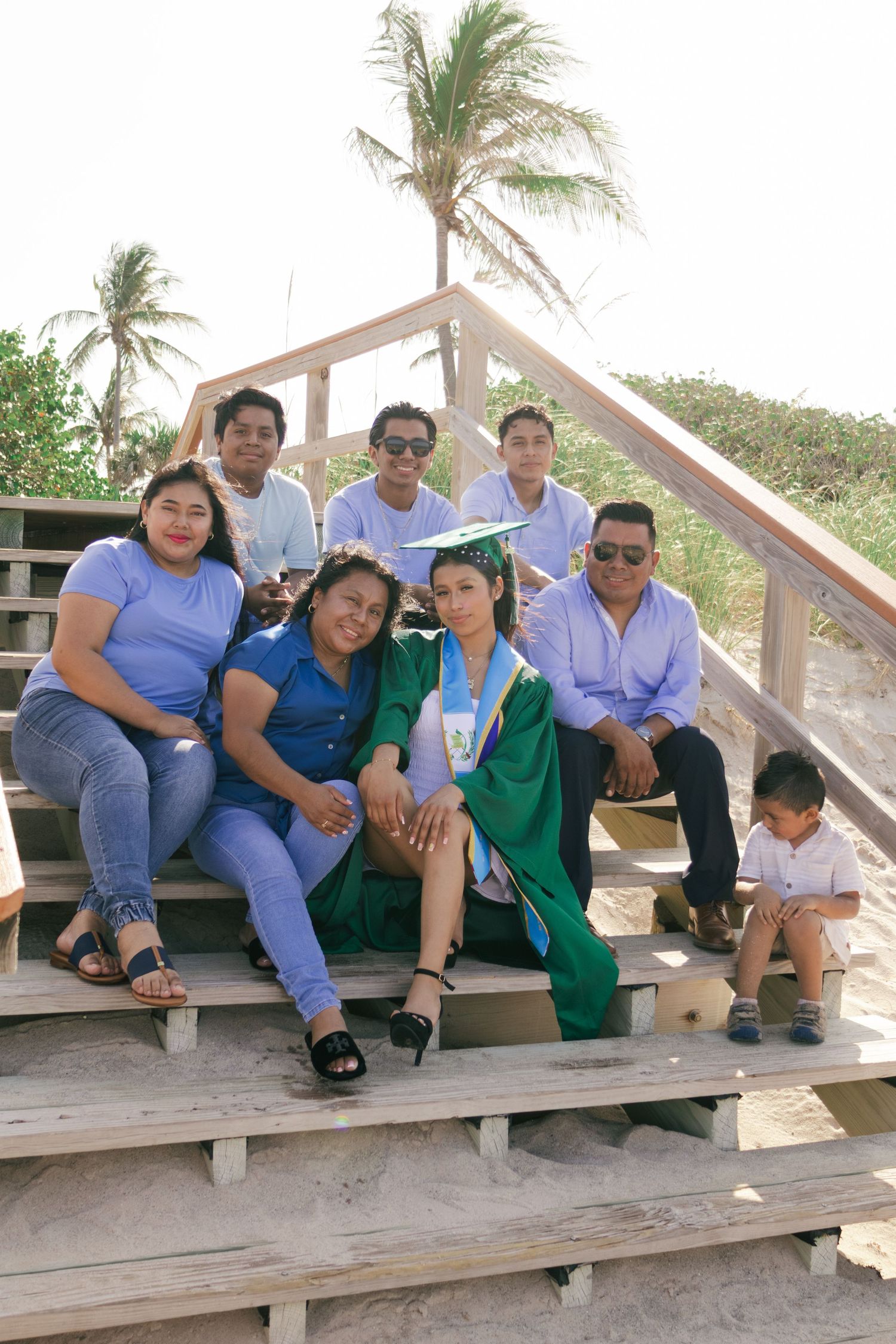 Family group photo on beach boardwalk steps celebrating graduation, graduate in green cap and gown surrounded by loved ones.