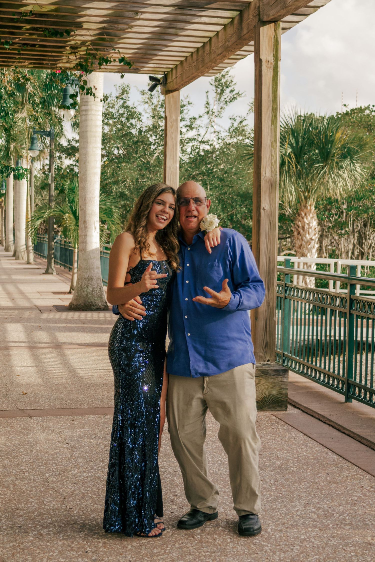 A woman in a blue sequin gown poses with an older man in a blue shirt under a pergola.