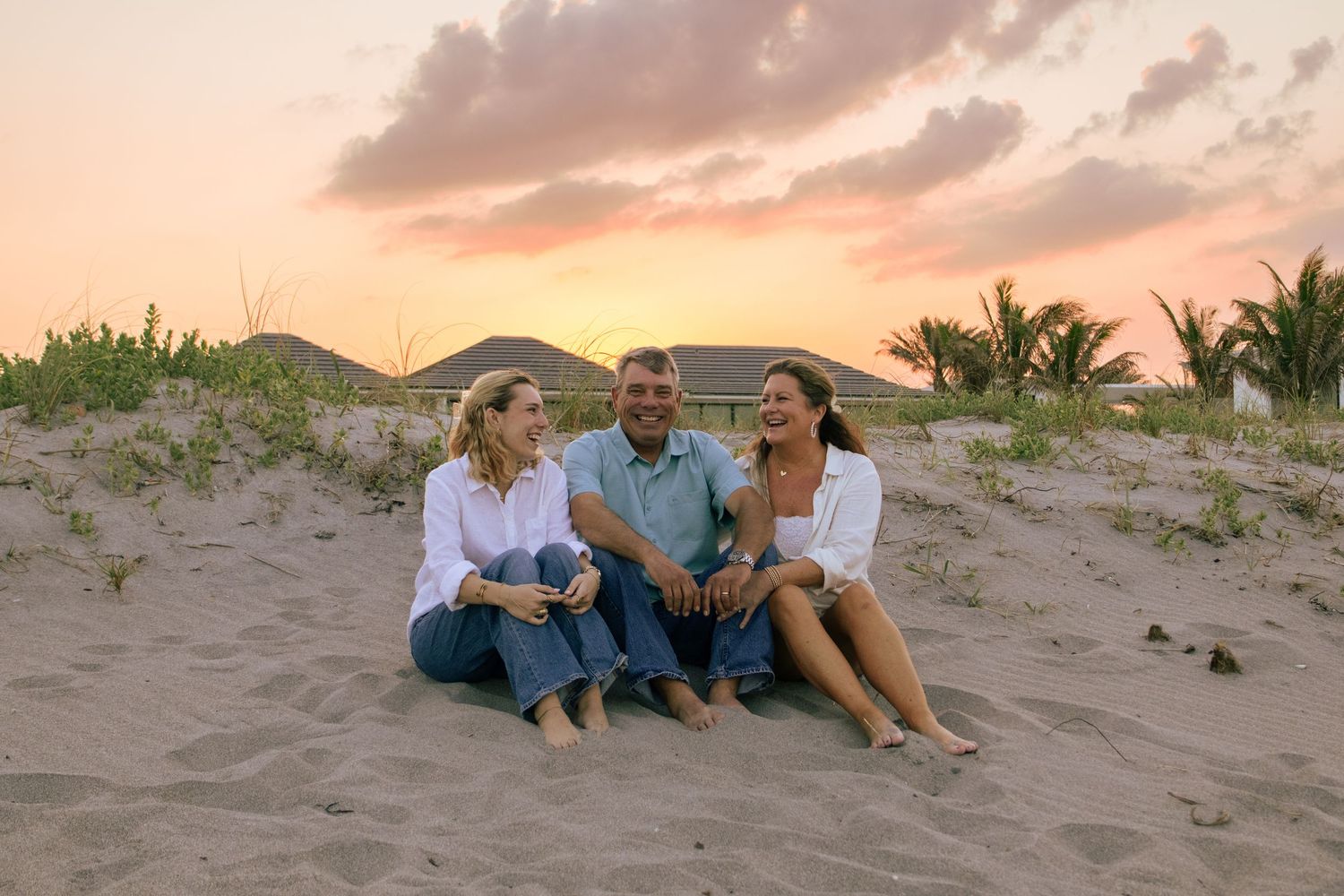 Three friends smiling and sitting together on sandy beach at sunset with palm trees and colorful sky.