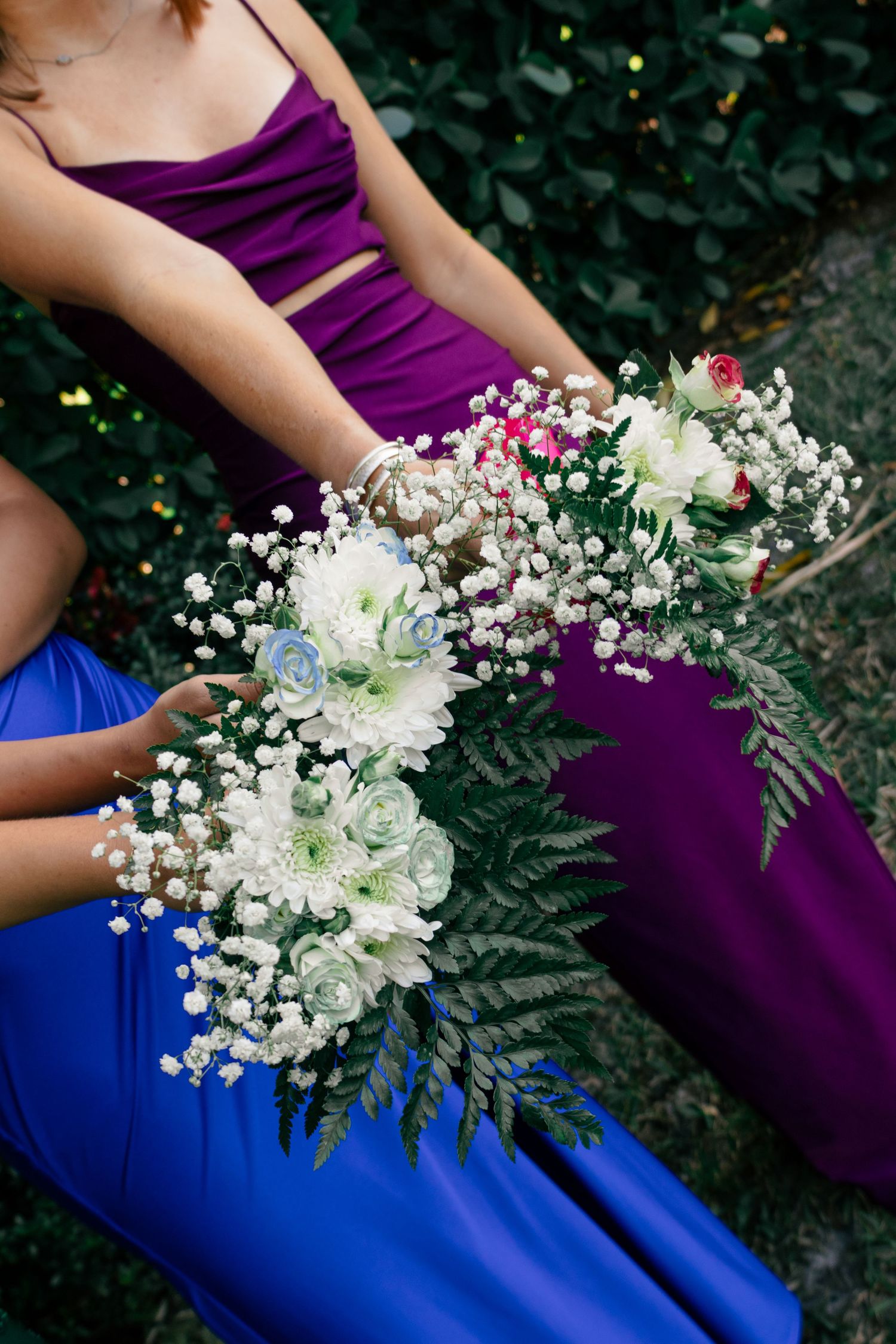 Close-up of two prom attendees in blue and purple gowns holding white floral bouquets with ferns.