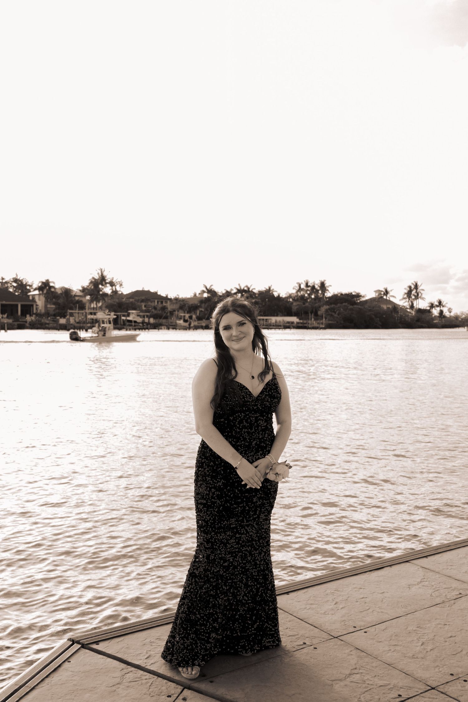 A sepia-toned portrait of a teenage girl in a sequin prom dress posing on a scenic Florida waterfront dock.