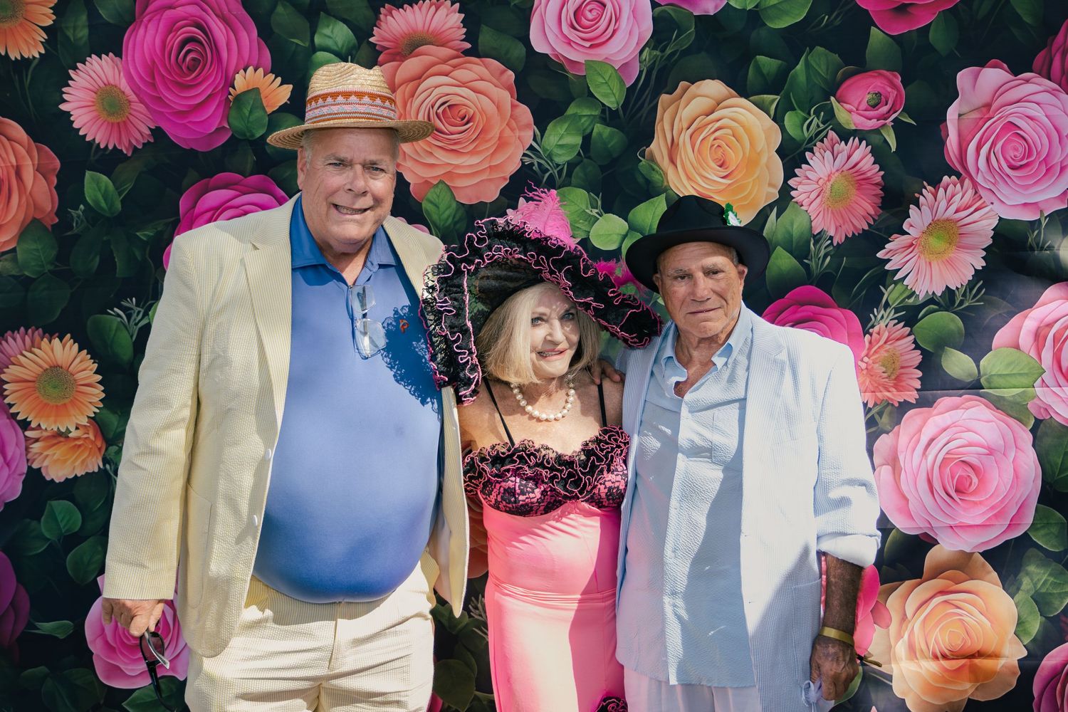 Three guests posing and smiling in front of a vibrant floral backdrop at a Kentucky Derby party.