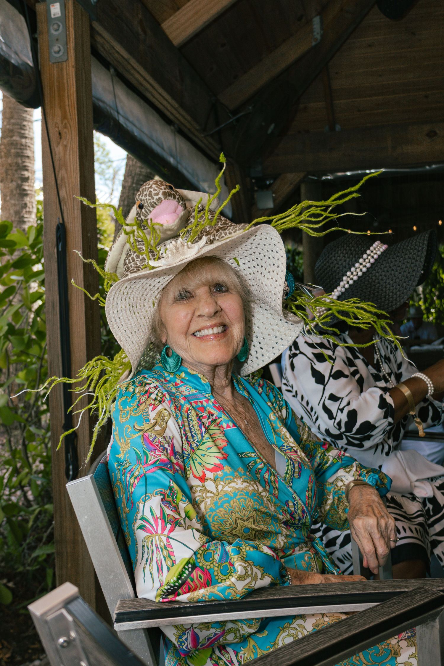 Elderly woman smiling in colorful outfit and white hat with green floral decorations.