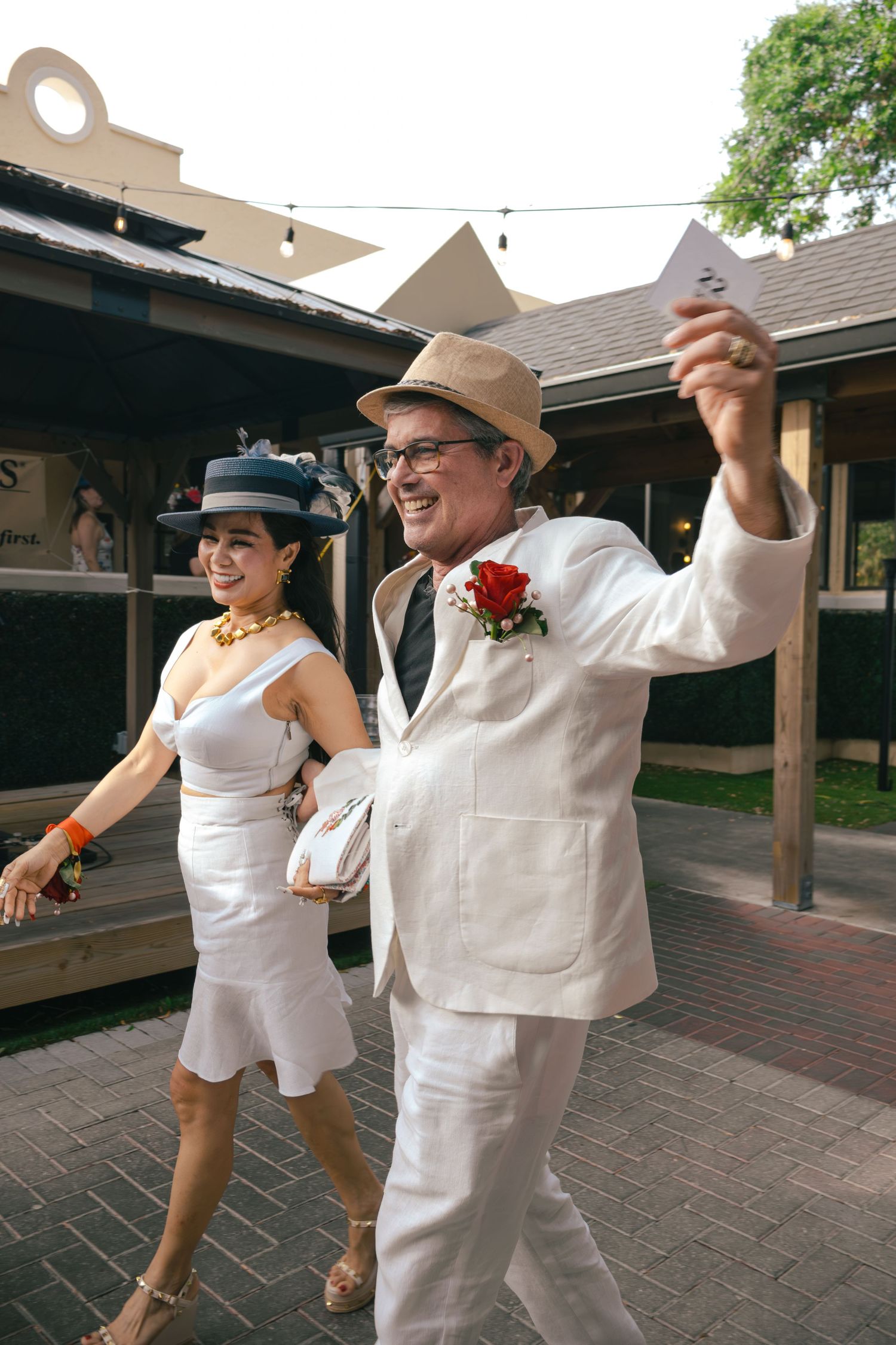 Smiling couple in white outfits walking at outdoor event, man raising paper triumphantly.
