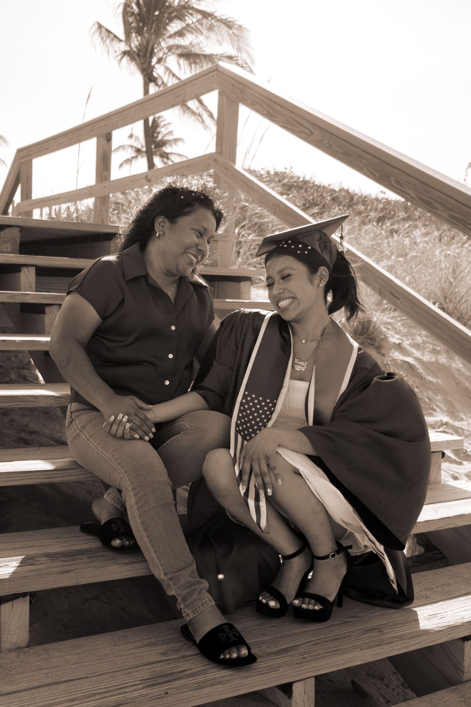 Sepia-toned image of mother and daughter in graduation attire sharing a joyful moment on beach steps.