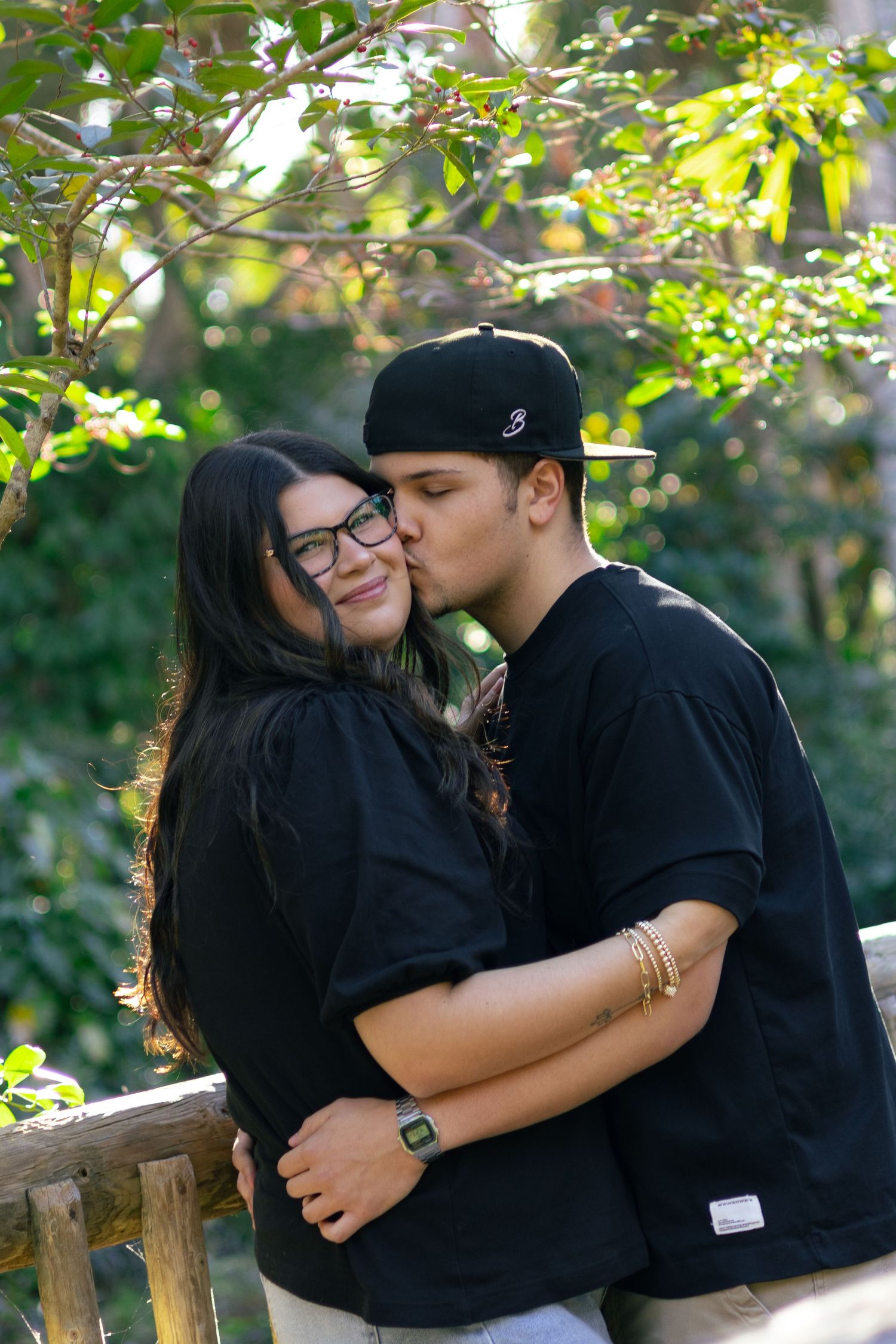 Young couple in black outfits sharing a kiss near a wooden fence surrounded by green trees.