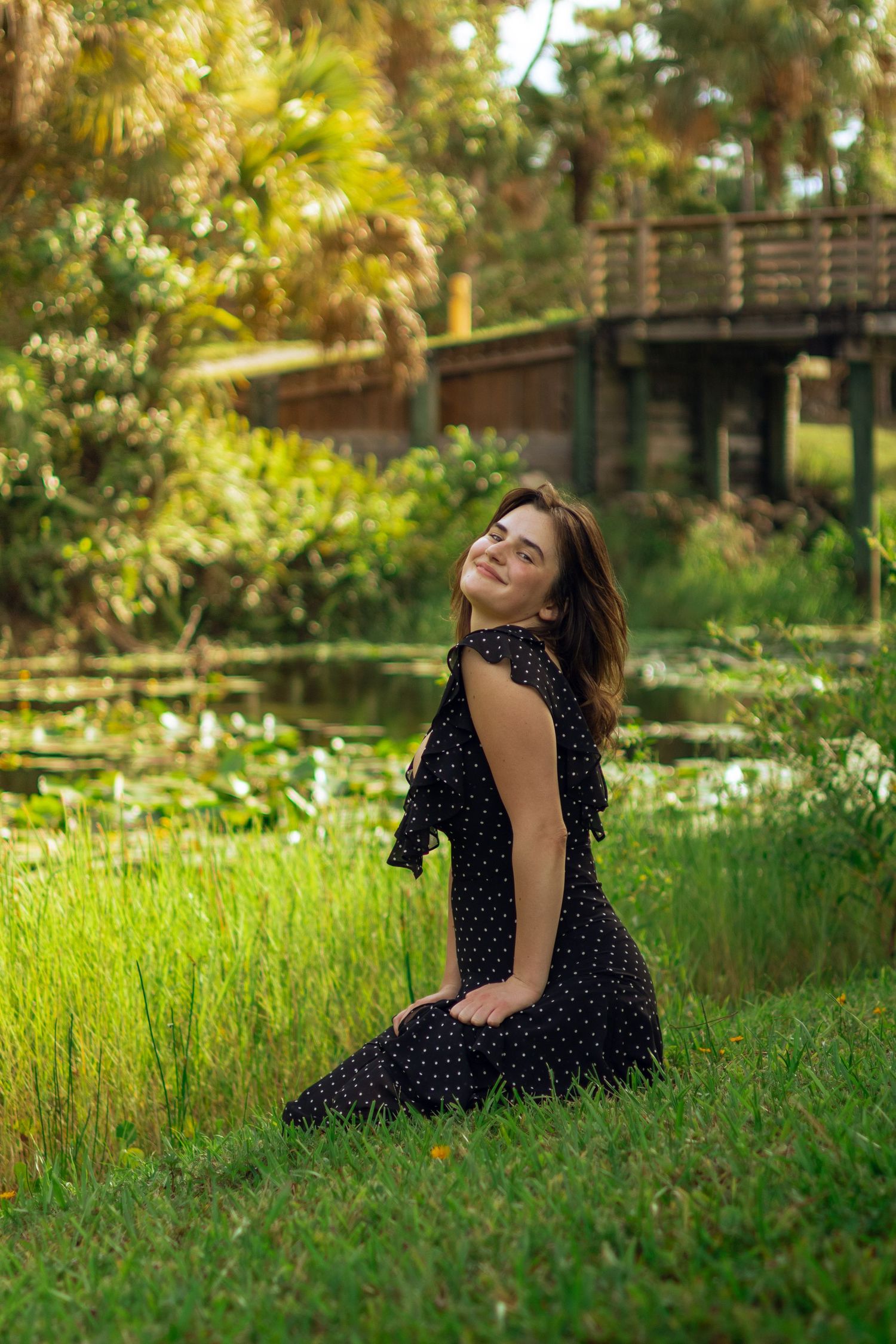 Smiling woman in black polka dot dress kneeling on grass by a serene pond in lush park.