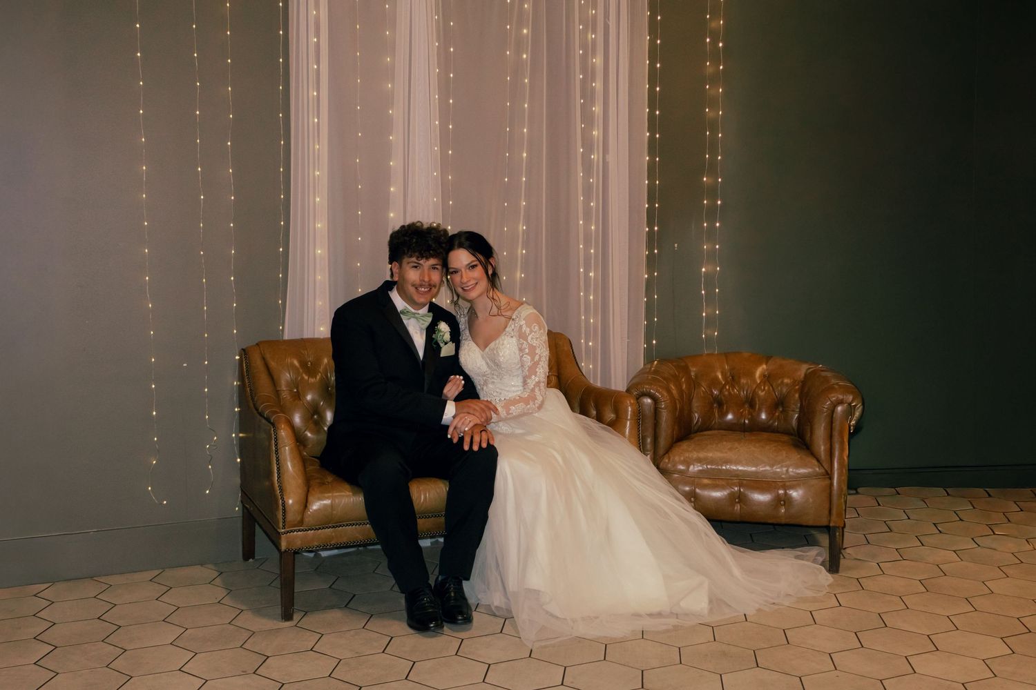 Bride and groom sit together on leather chairs in front of a twinkling fairy light backdrop at their wedding reception.