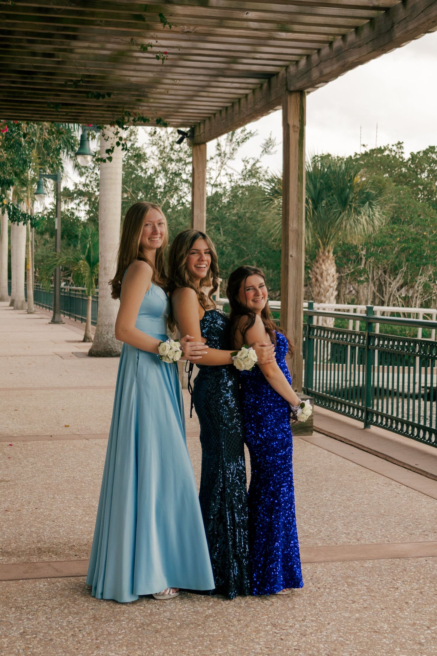 Three teenage girls in blue prom dresses pose under a pergola walkway with white floral corsages.