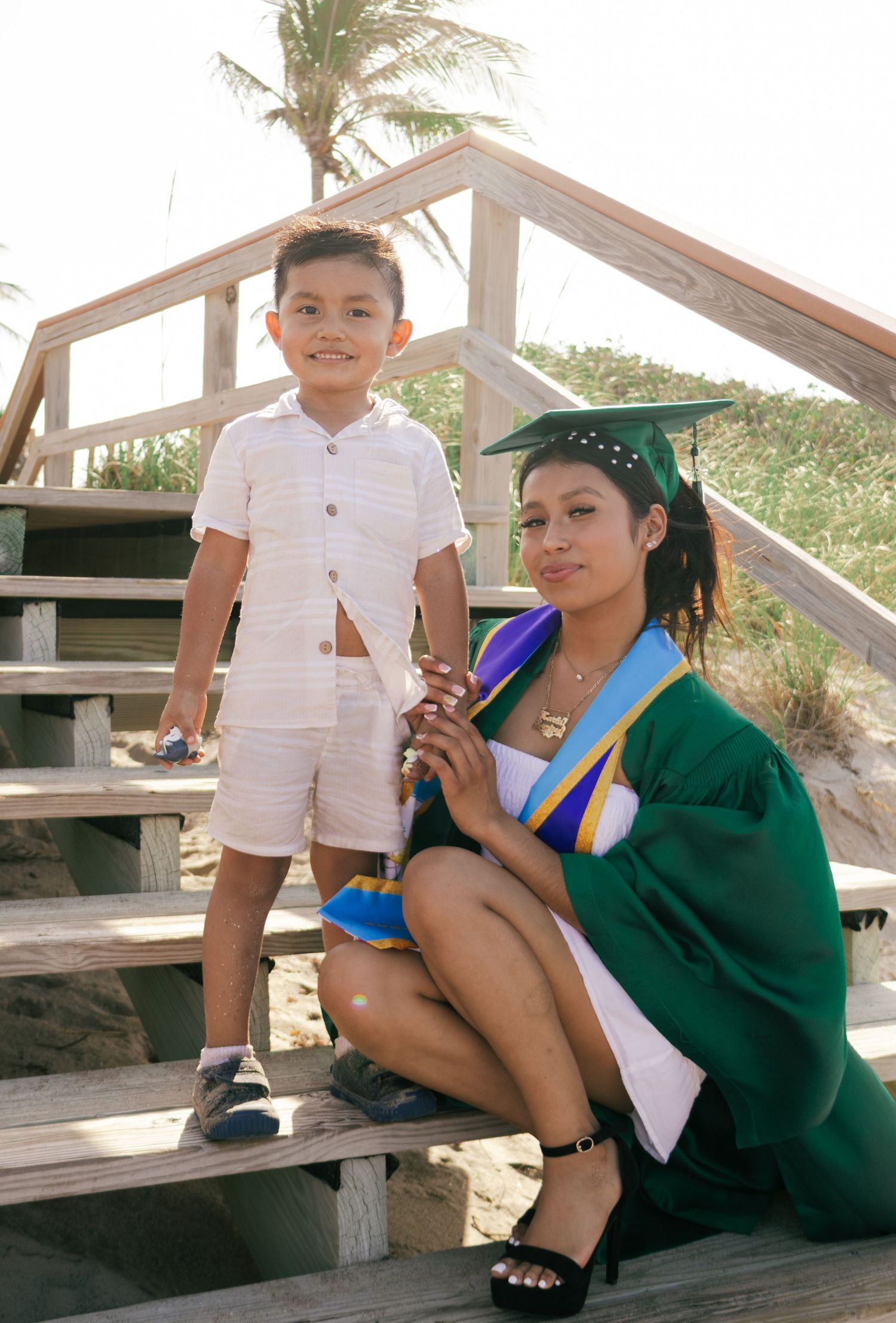 A female graduate in green cap and gown poses with a young boy on wooden beach boardwalk stairs.