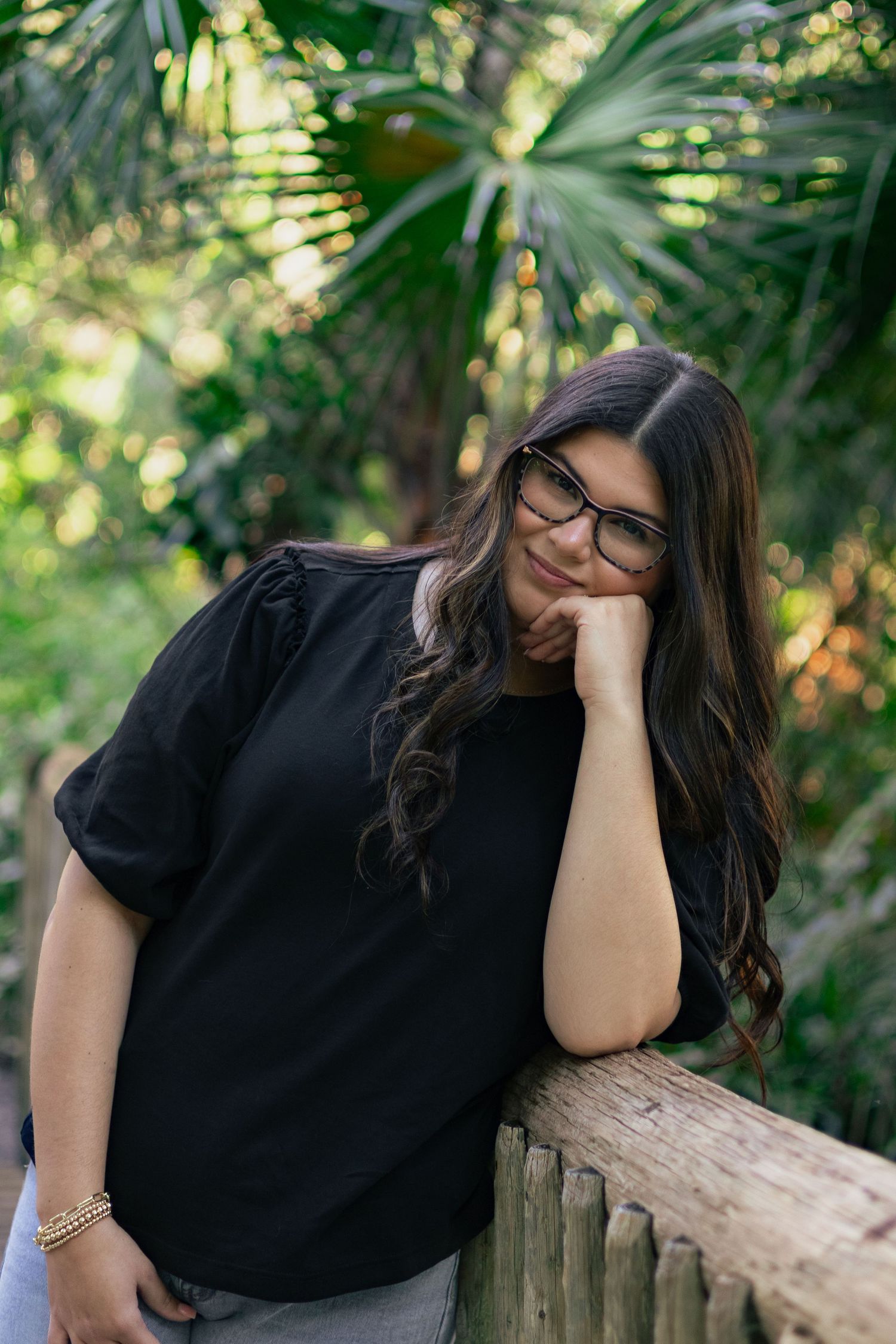 Young woman with glasses and long dark hair in black top leaning on wooden fence in lush garden.