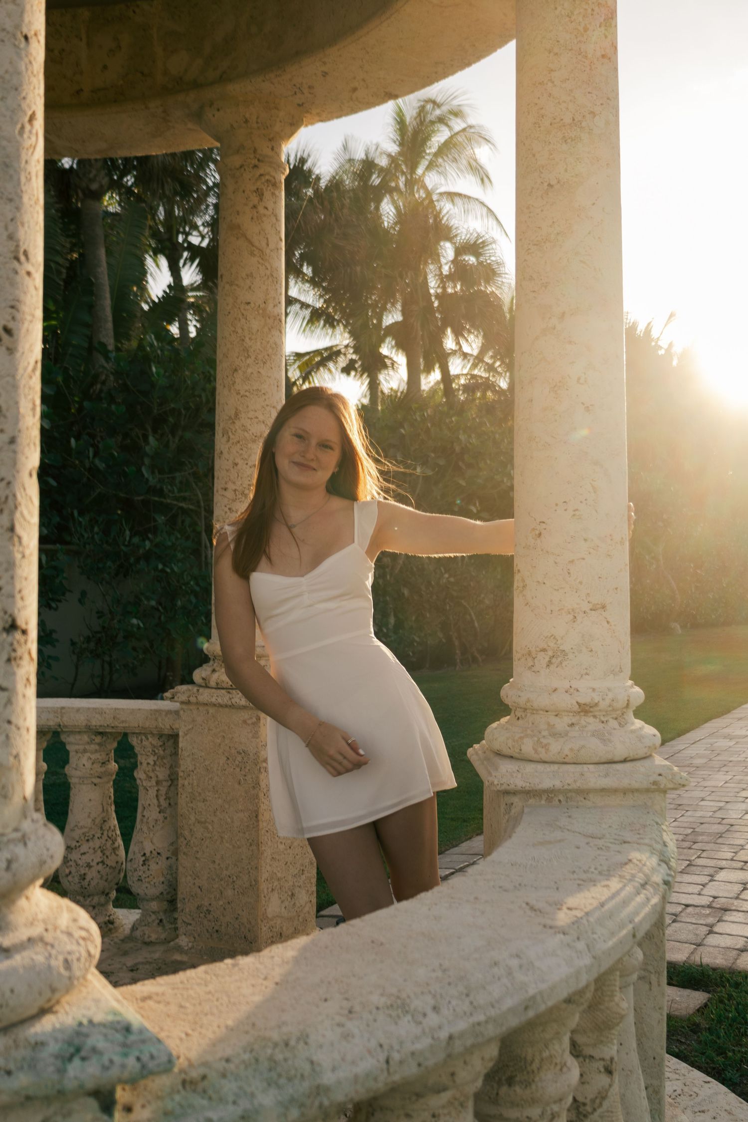 Young woman in white dress leaning on stone column in classical rotunda with golden sunset light.