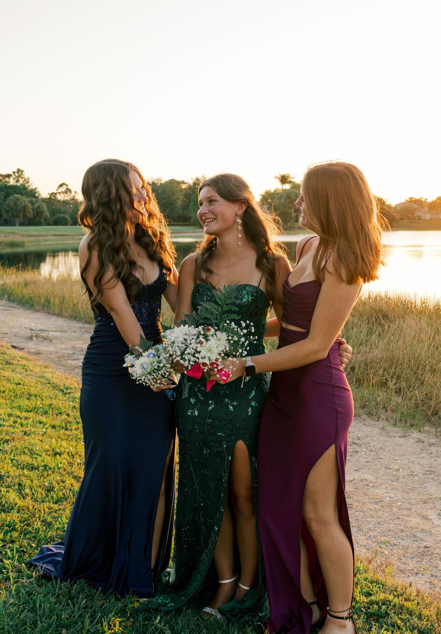 Three young women in elegant navy, green sequin, and burgundy prom gowns pose by a lake at golden hour.