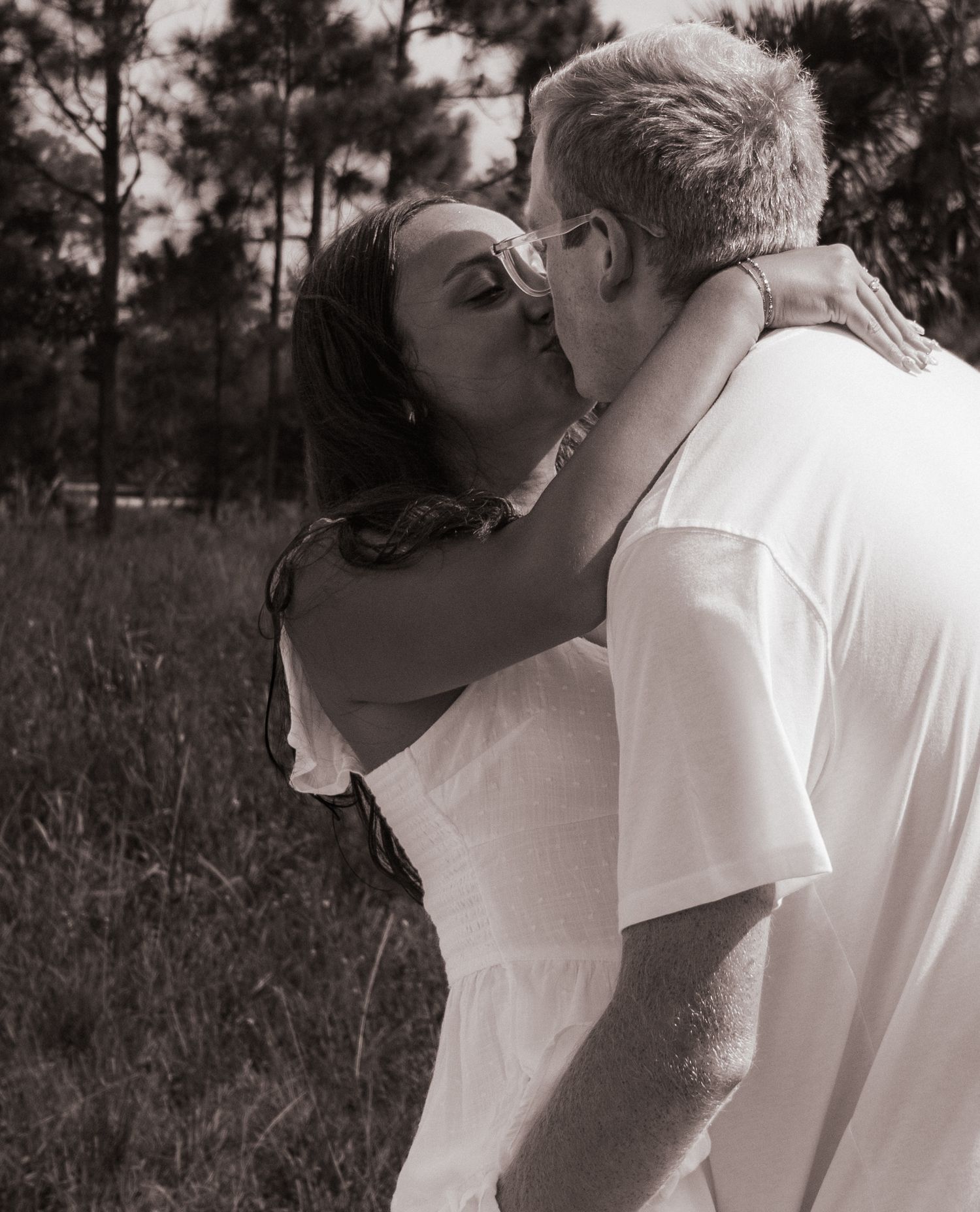 Black and white romantic photo of couple sharing a kiss outdoors among pine trees.