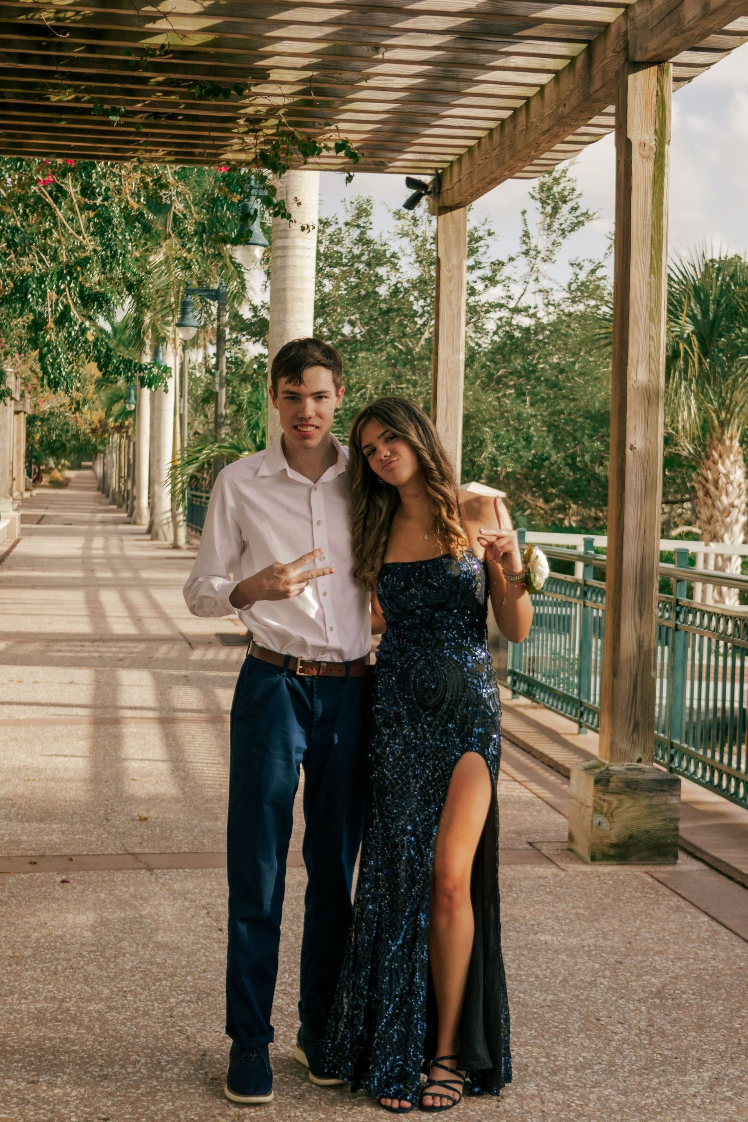 Young couple in formal attire posing together under a vine-covered pergola at an outdoor venue.