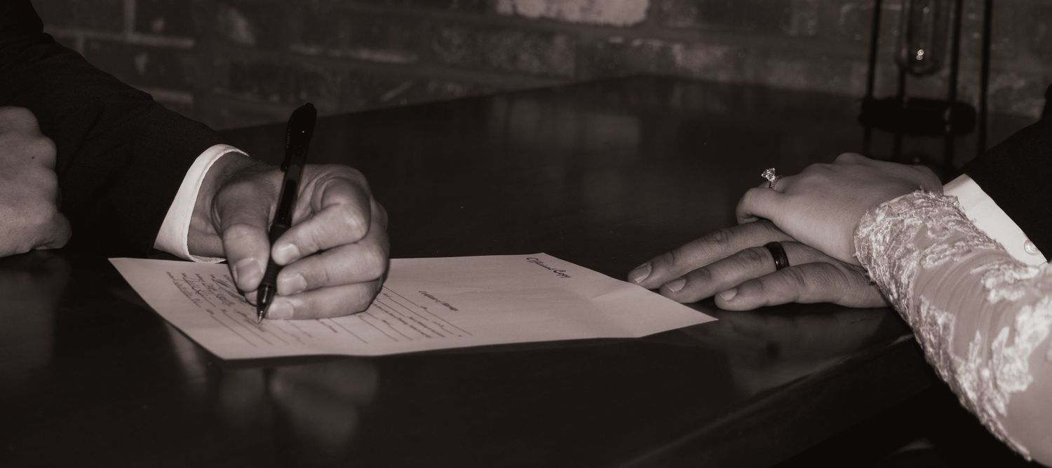 Black and white photo of groom signing wedding certificate with bride beside him.