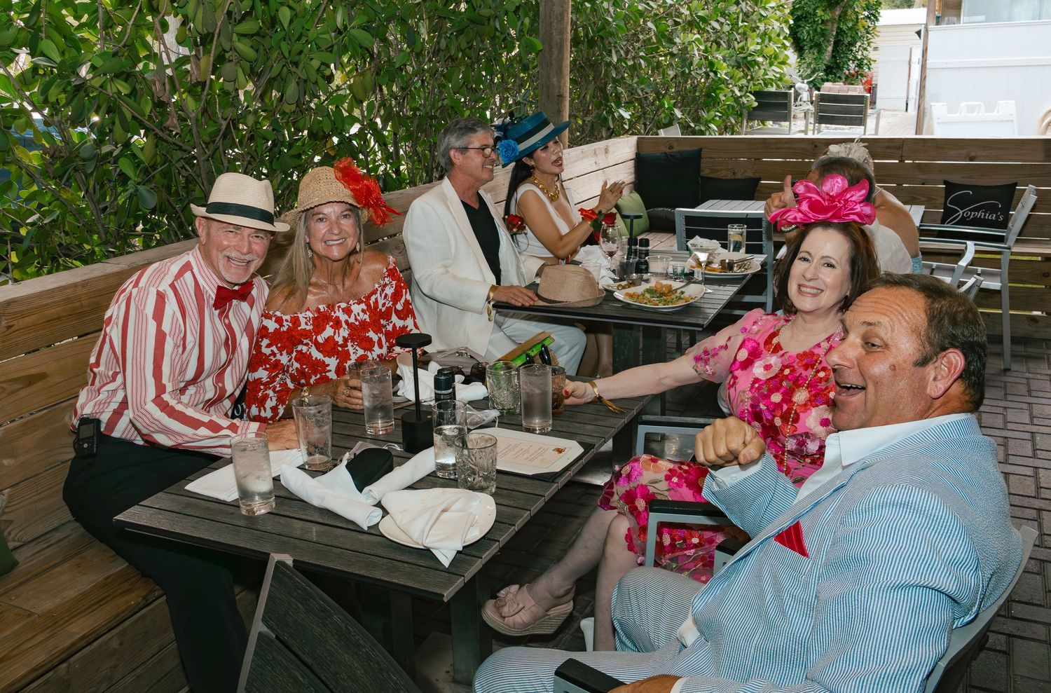 Cheerful group of friends in colorful Kentucky Derby attire dine together at an outdoor restaurant.