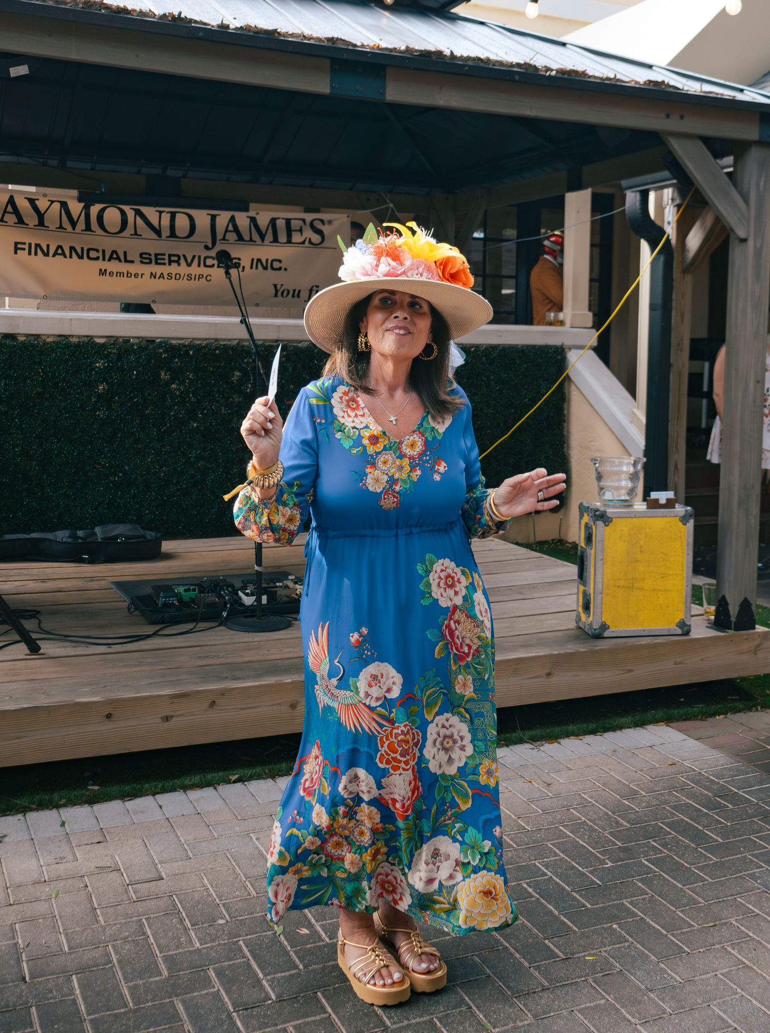 Woman in floral blue dress and decorated hat poses at outdoor Kentucky Derby party near Raymond James stage.
