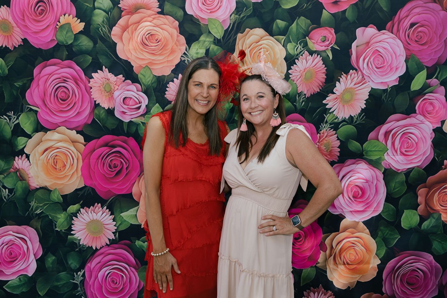 Two women in red and white dresses pose in front of a colorful floral backdrop with pink and orange roses.