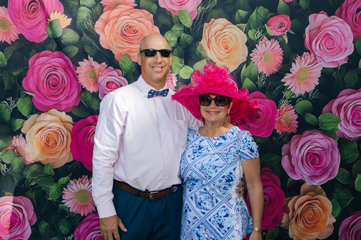 Couple posing in front of a floral backdrop, woman wearing a pink Kentucky Derby hat.