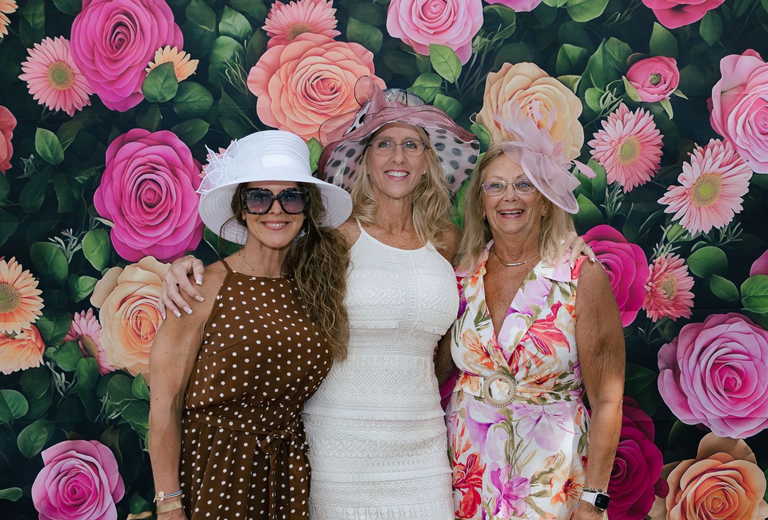 Three women in Derby hats posing before vibrant pink and orange floral photo backdrop.