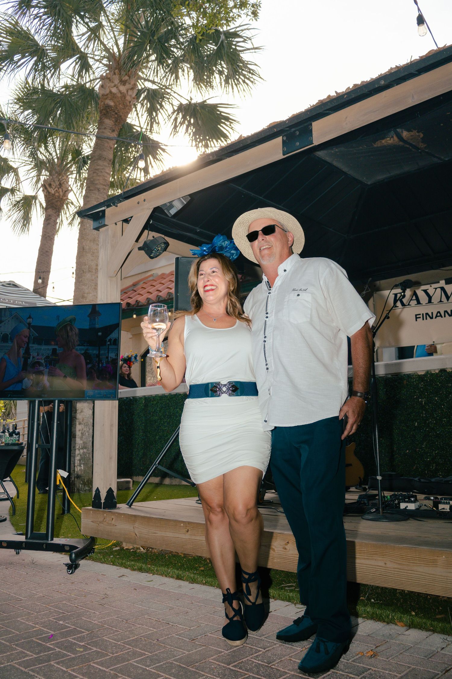 Couple in white outfits and festive hats toasting with champagne at an outdoor Kentucky Derby party.