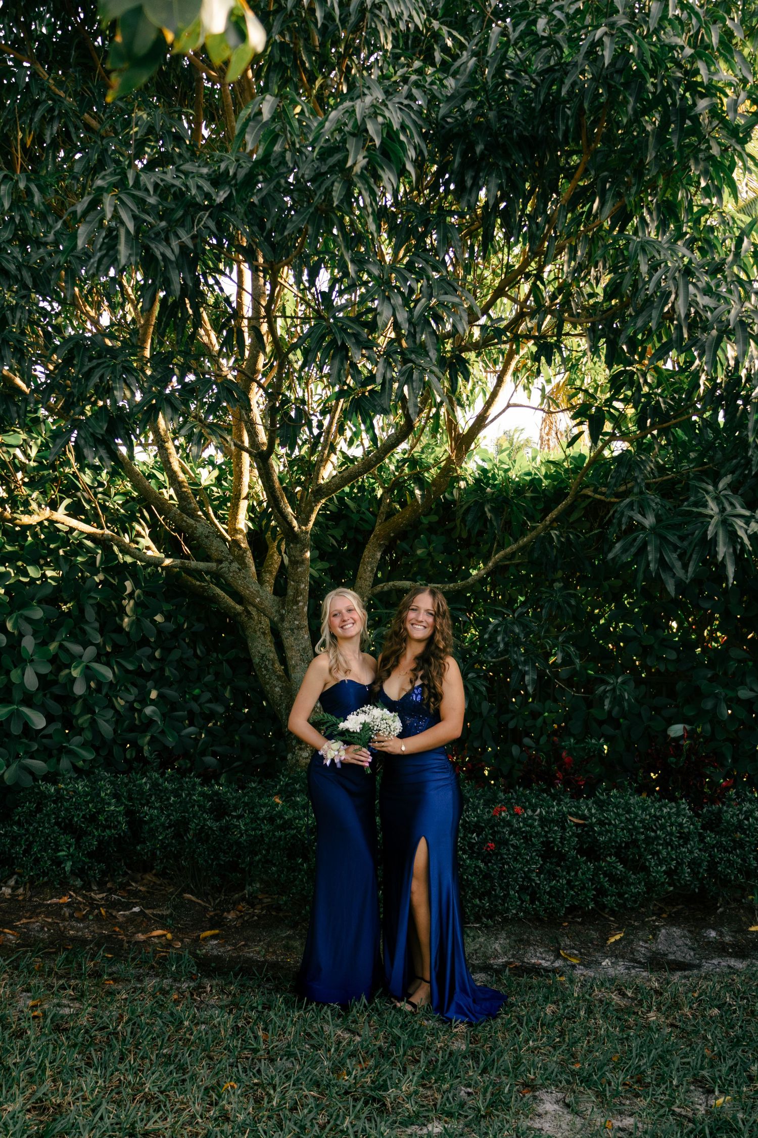 Two bridesmaids in navy blue gowns holding white bouquets standing under large green tree.