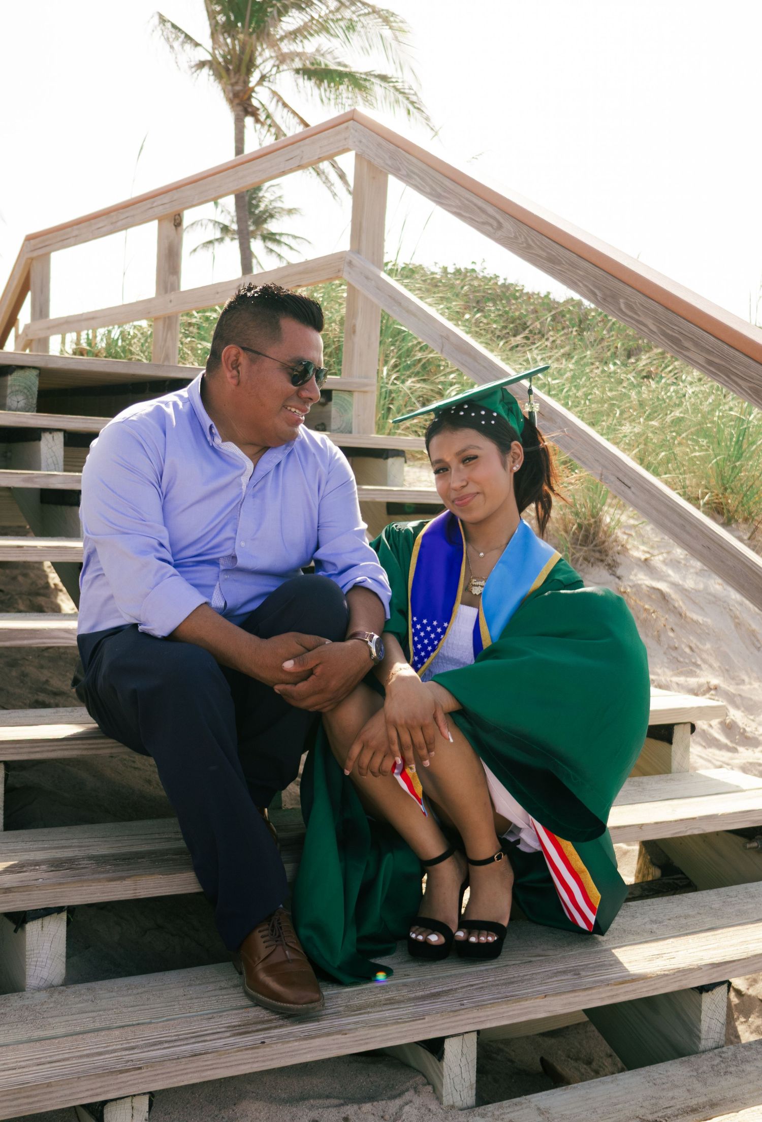 Graduate in green cap and gown sitting on wooden beach stairs sharing a quiet moment with proud father.