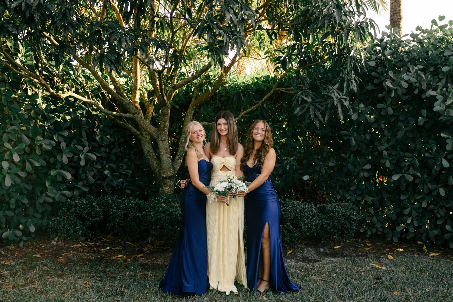 Three bridesmaids pose outdoors, two in navy blue gowns flanking one in a champagne dress, holding bouquets.